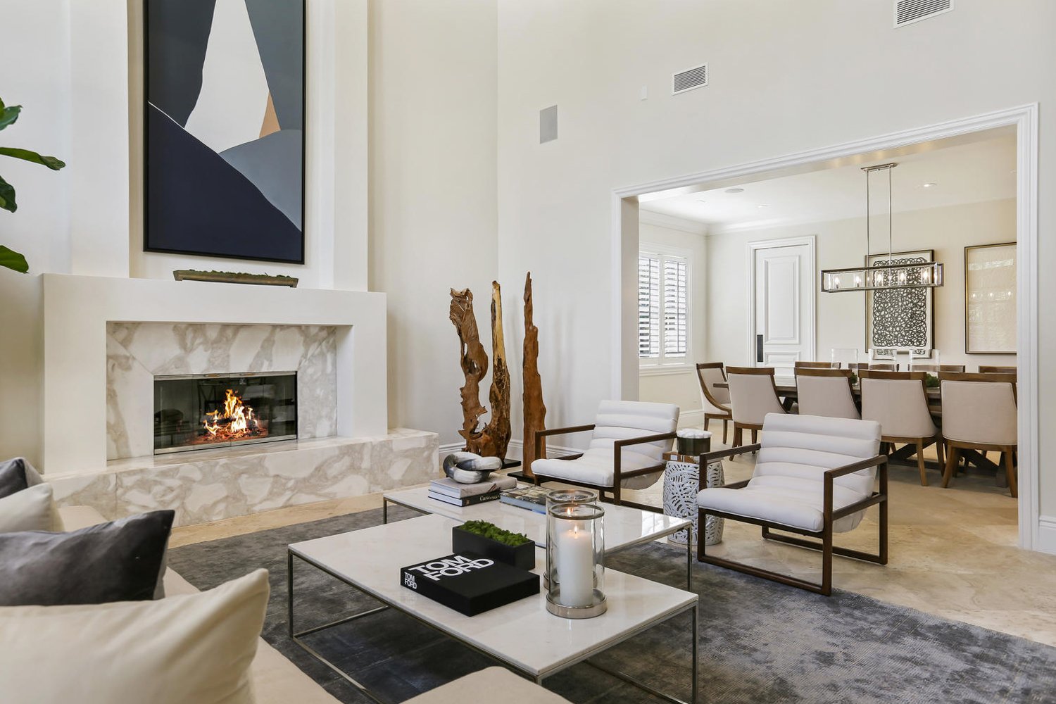 Listing photo of a home in Pinecrest. Showing a modern living room with a fireplace, abstract wall art, two white armchairs, coffee table with books and candles, and a dining area with a wooden table and chairs in the background.