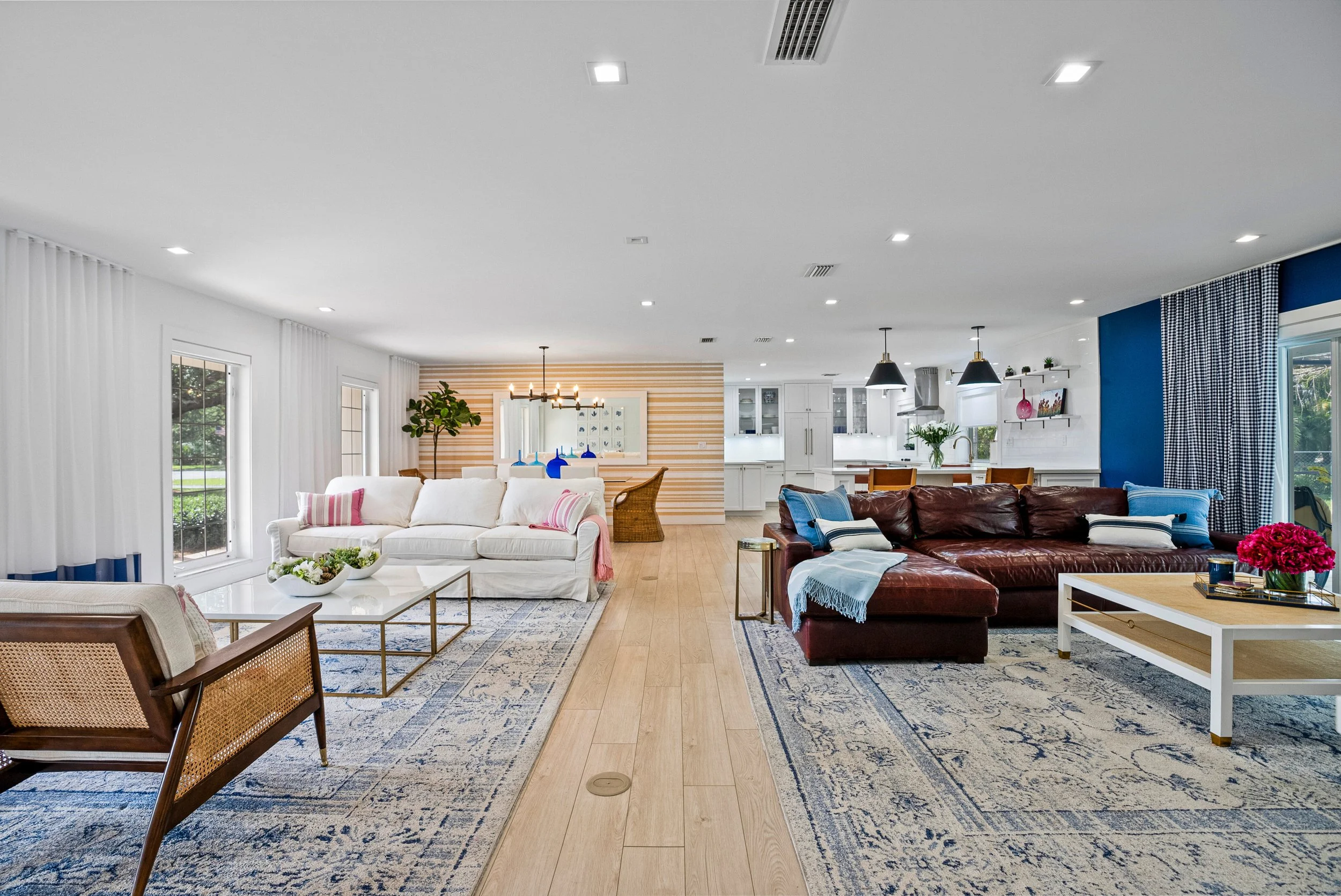 Spacious modern living room with white and blue walls, large windows with white curtains, a white sofa with pink and white pillows, a leather sofa with blue and white pillows, a wooden coffee table with a floral arrangement, and a patterned blue and beige rug. The open kitchen is visible in the background with white cabinets, a dining table, and black pendant lights.