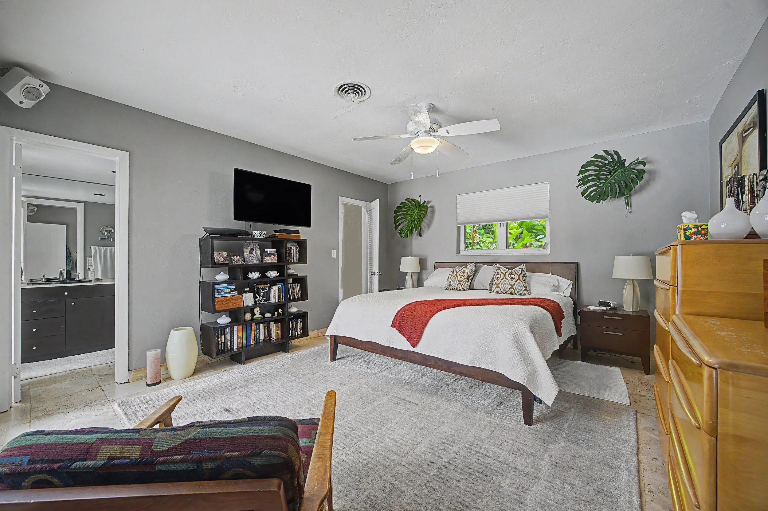 A Coral Gables bedroom with a bed, a dresser, a television, a bookshelf, and decorative vases, with a window and ceiling fan.