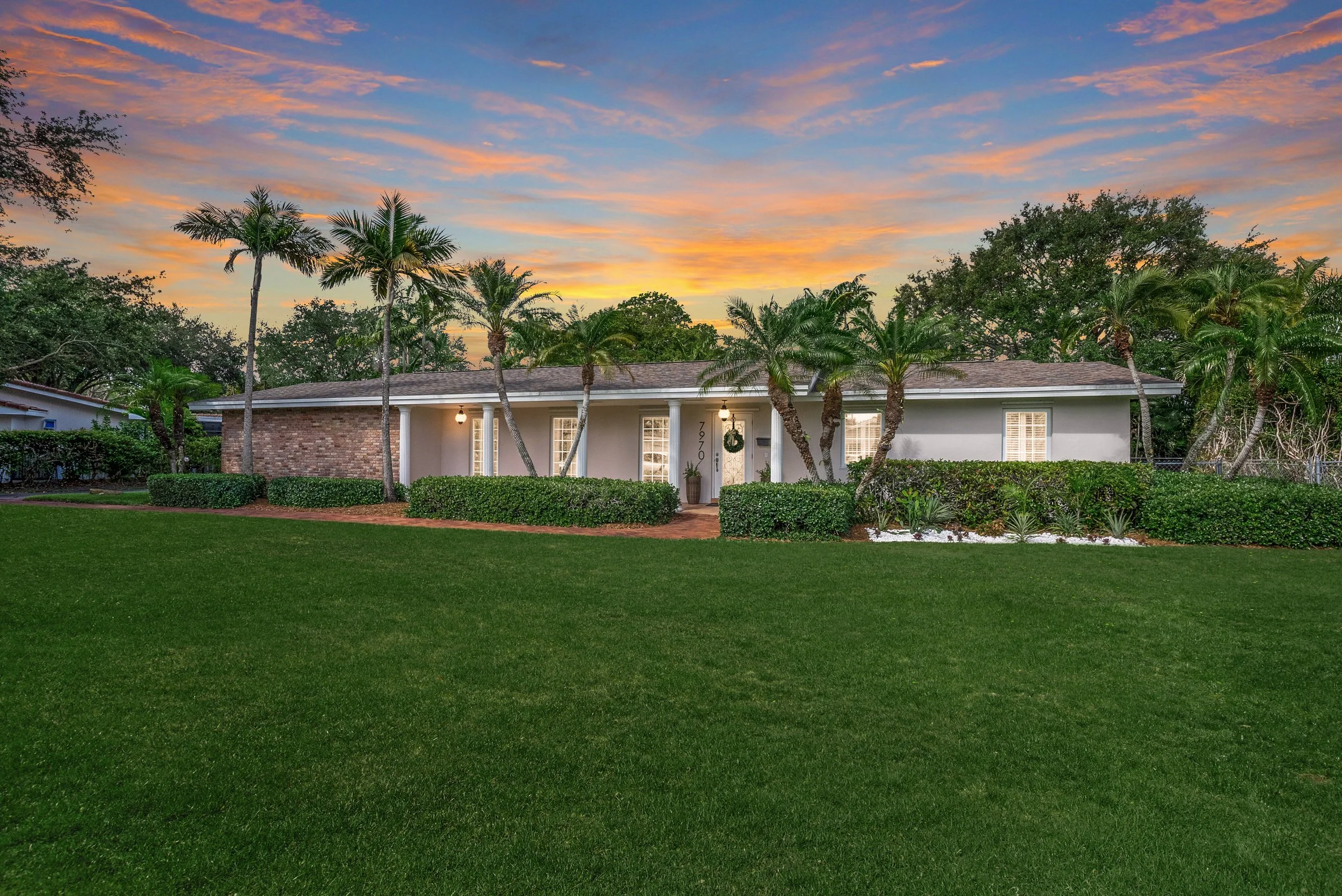 A listing photo of a suburban house with a well-maintained lawn and palm trees in front, under a colorful sunset sky with orange, pink, and blue clouds.