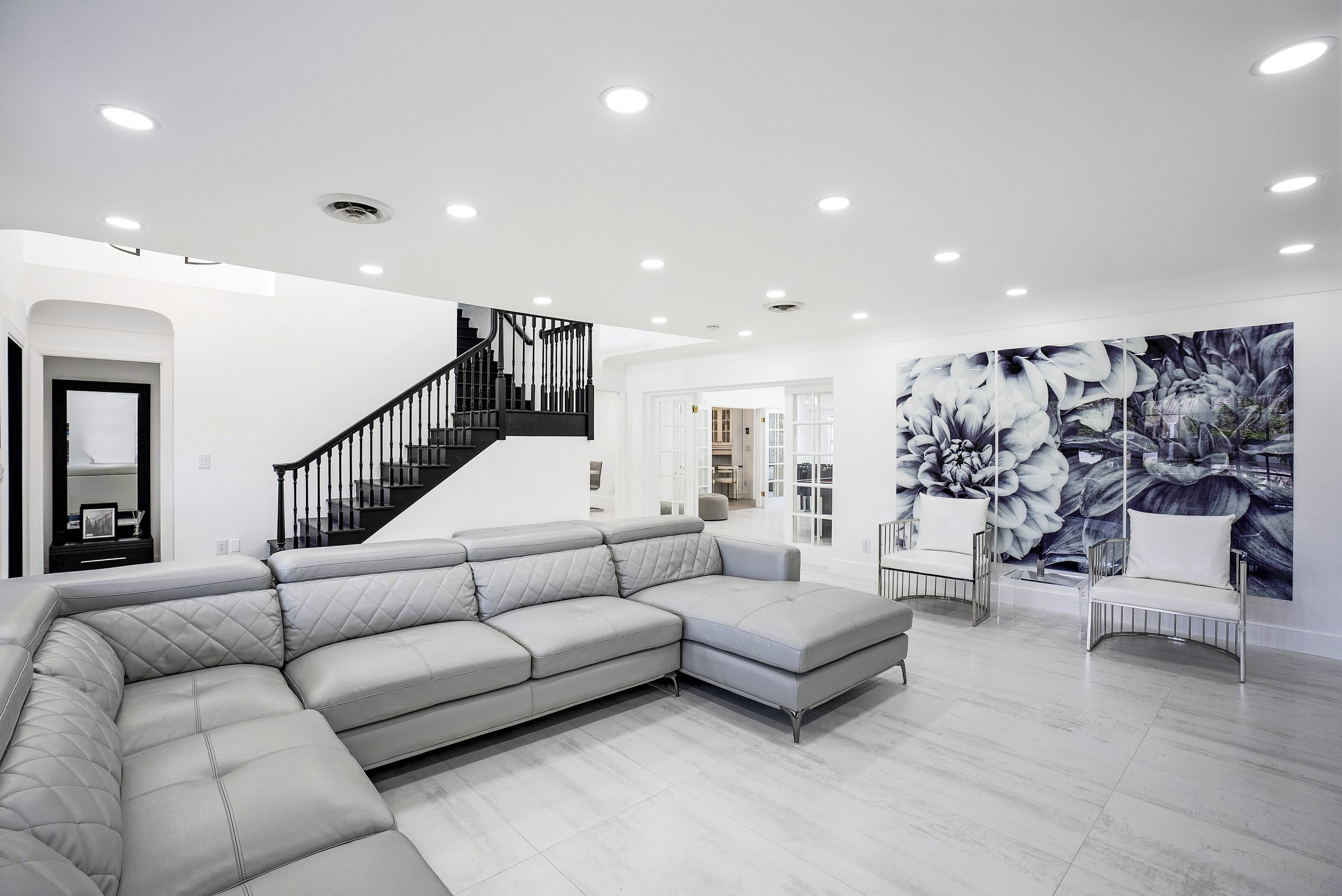 Professional real estate photography of a luxury living room in Fort Lauderdale, Florida, featuring a gray leather sectional sofa, elegant black staircase, white tile flooring, modern accent chairs, and large floral wall art.