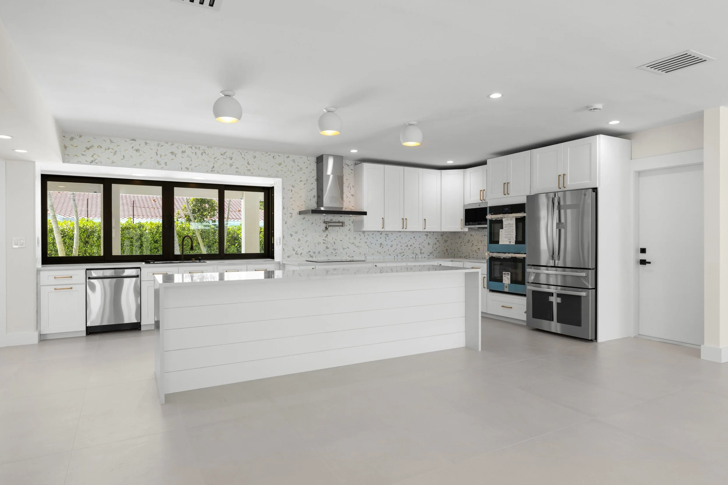 Modern kitchen with white cabinets, stainless steel appliances, and large window overlooking greenery.