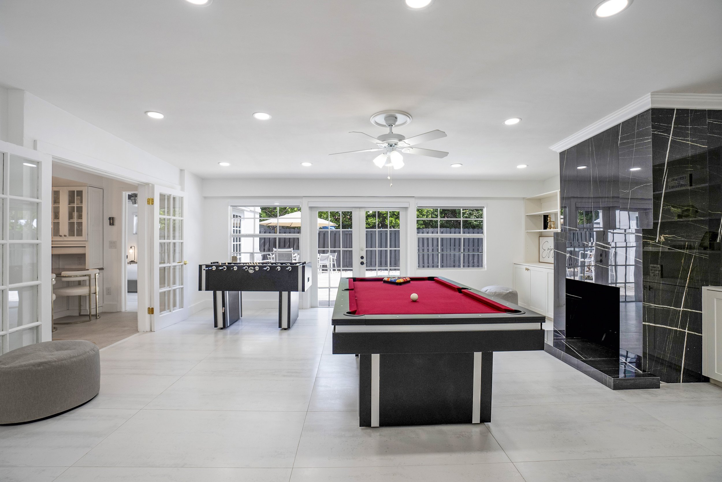 Professional real estate photography of a spacious luxury home game room in Fort Lauderdale, Florida, featuring a red-felt pool table, foosball table, black marble fireplace, white tile floors, and French doors overlooking the patio.
