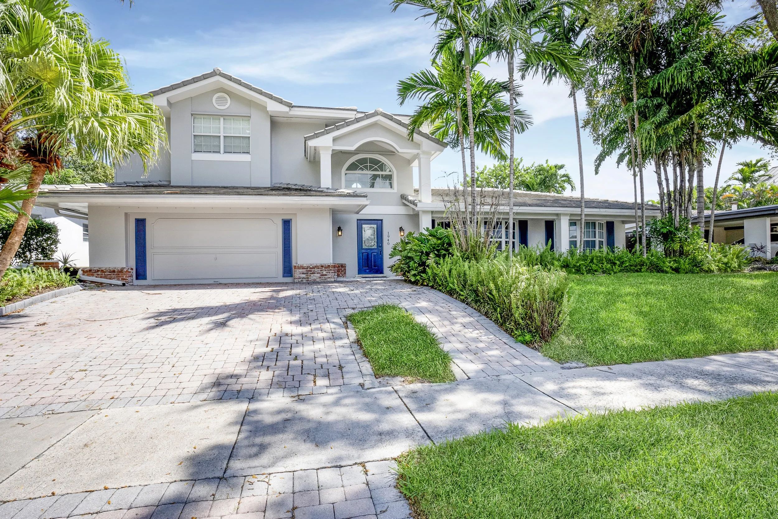 Professional real estate photography of a luxury two-story home in Fort Lauderdale, Florida, featuring tropical landscaping, palm trees, a circular paver driveway, and a white exterior with blue accents.