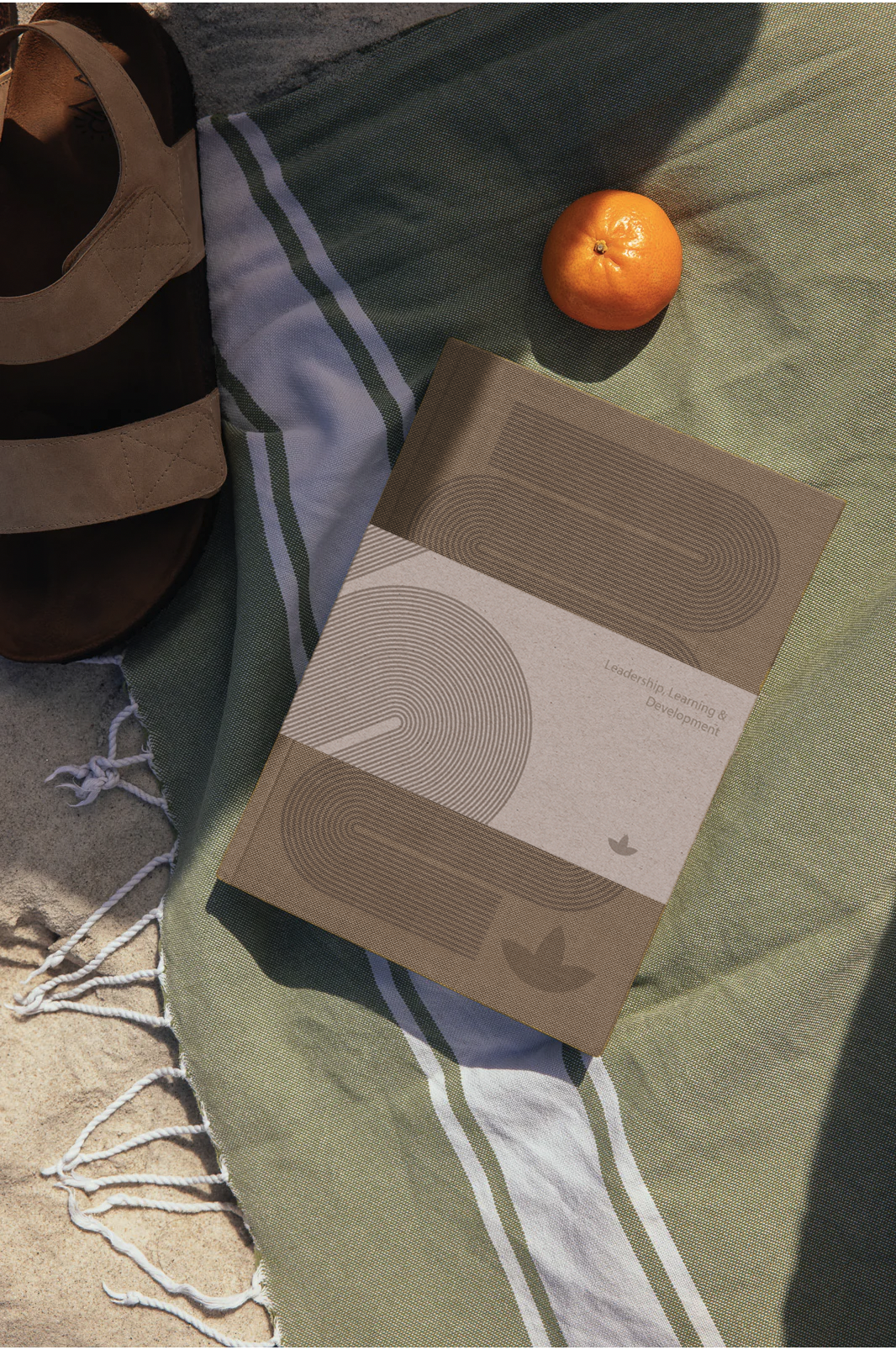 A sandy beach scene with a pair of tan sandals, a green and white striped beach towel, an orange, and a brochure titled 'Leadership Learning & Development'.