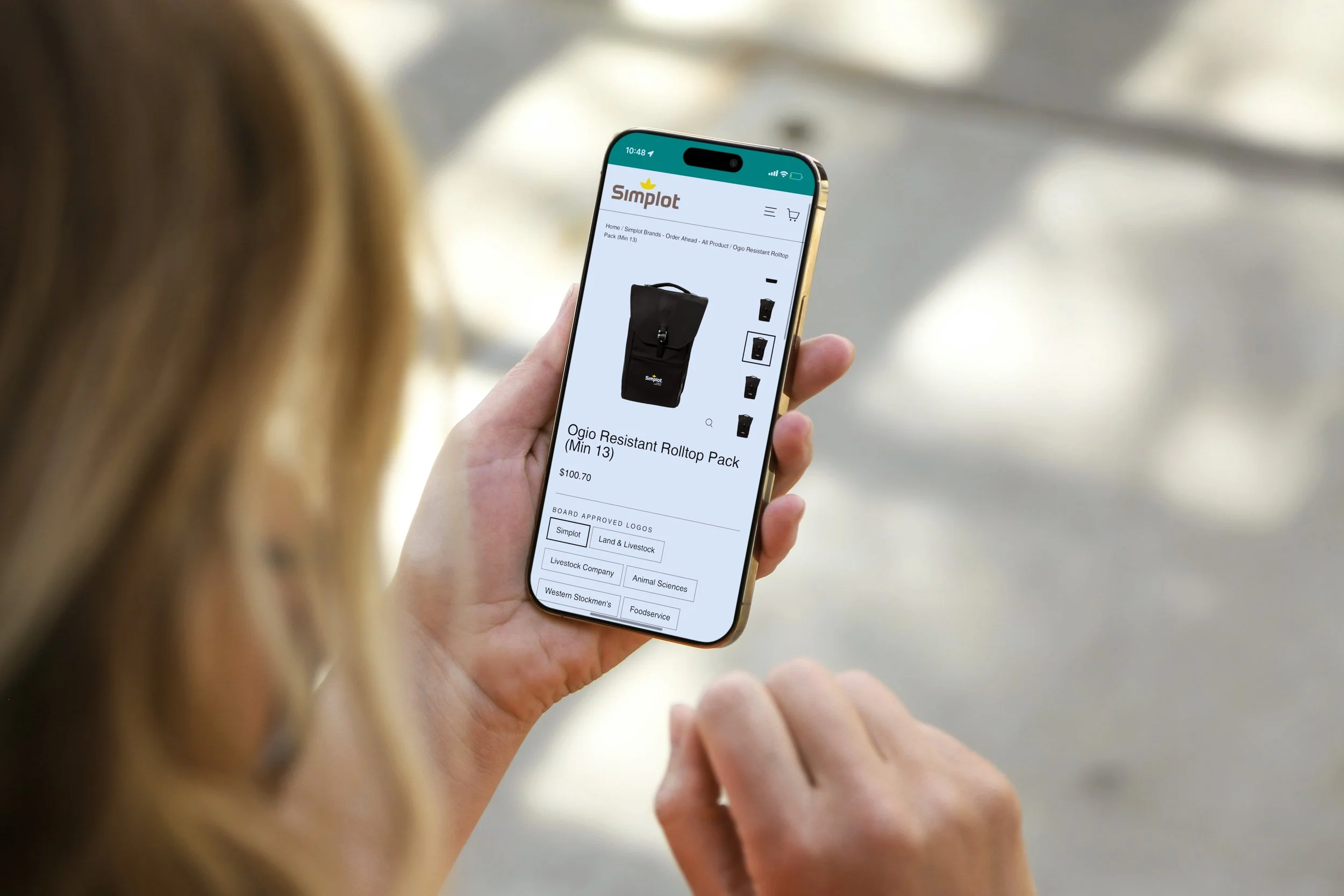 Person browsing on a smartphone in a marketplace or store, viewing a product page for a black Ogio Resistant Rolltop Pack on an e-commerce website.