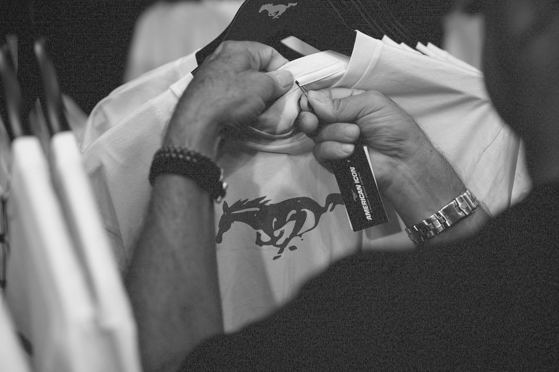 Close-up of hands sewing a white T-shirt with a Mustang logo, on a worktable, with a clothing tag reading 'American Icon' visible.