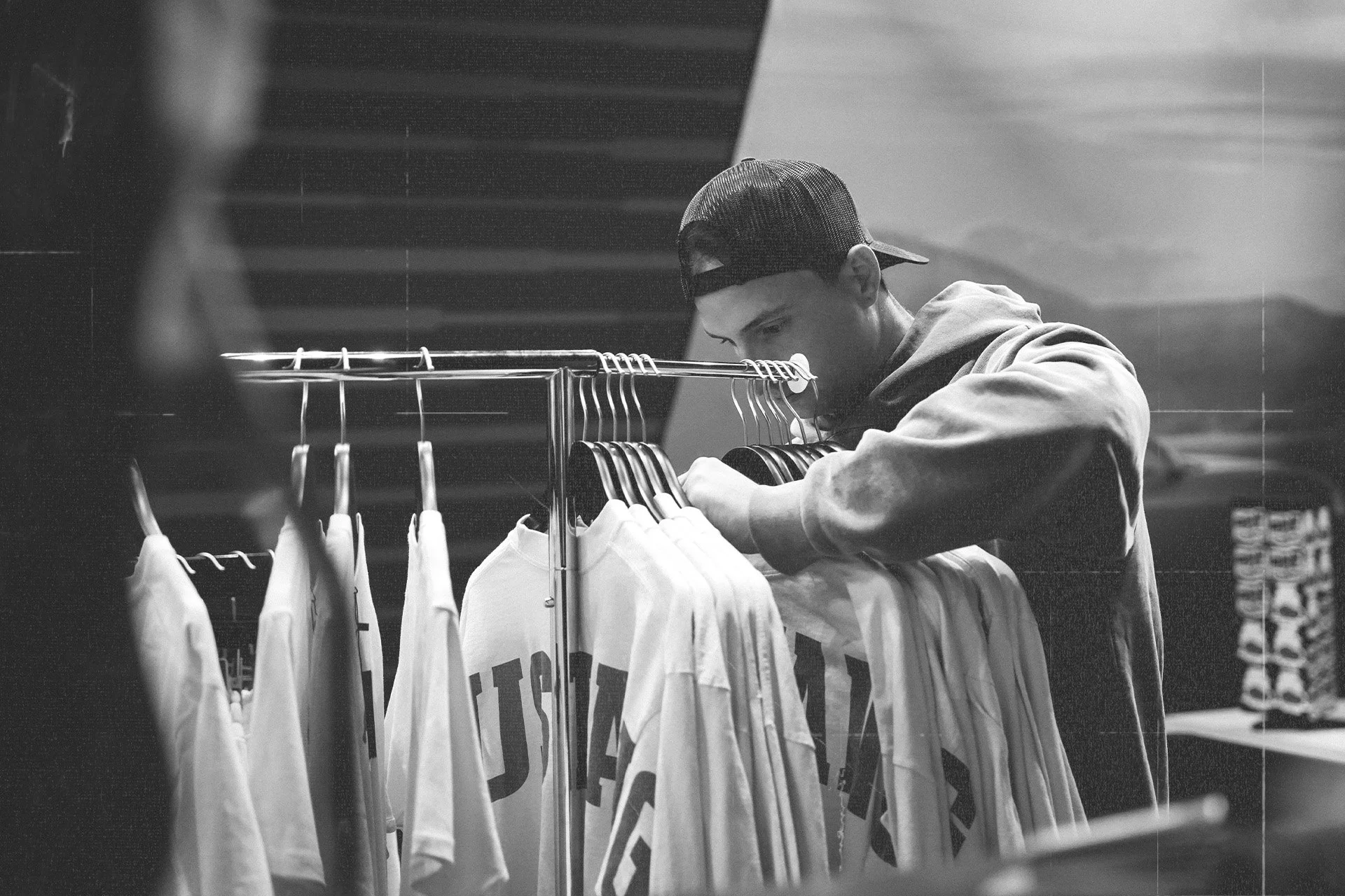 A young man wearing a backwards cap and hoodie shopping for t-shirts at a clothing store, with shirts on a rack in front of him.
