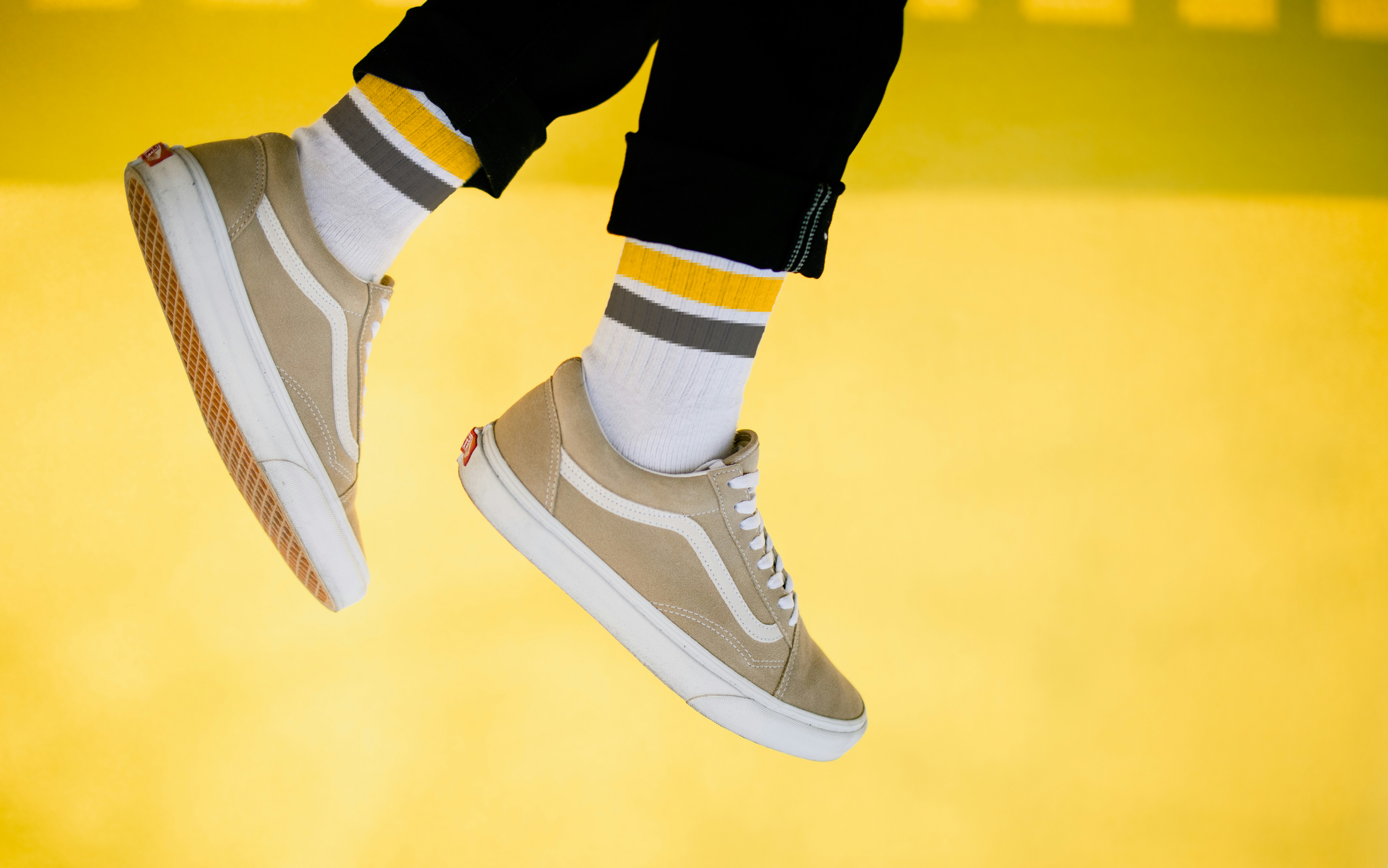 Close-up of a person wearing beige Vans sneakers, white socks with yellow, white, and gray stripes, and black pants against a yellow background.