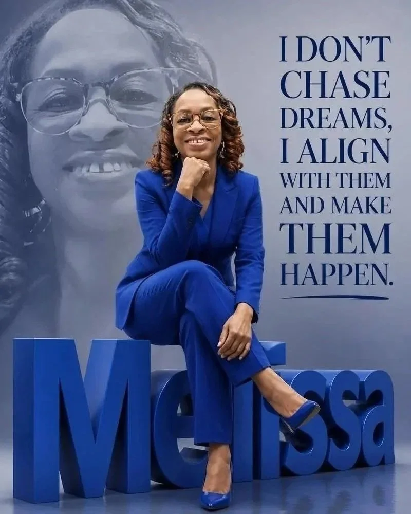 A woman in a blue suit sitting on large blue letters spelling 'MESA' at a booth or event with a large poster behind her. The poster features a large, faded black-and-white image of her smiling face and an inspirational quote: 'I don't chase dreams, I align with them and make them happen.'