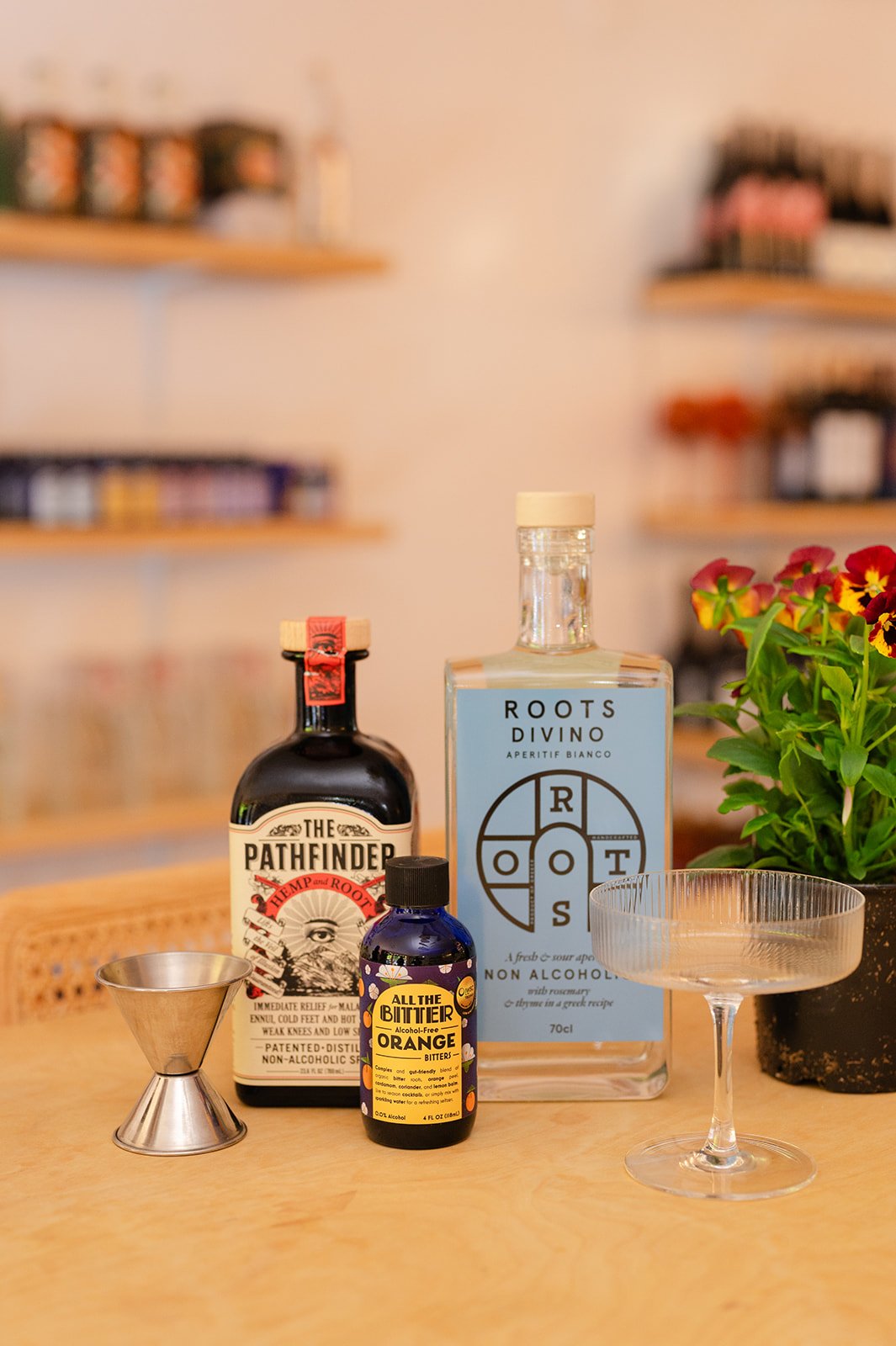 Assorted cocktail ingredients on a wooden surface, including a bottle labeled 'The Pathfinder,' a blue bottle labeled 'Roots Divino,' a small colorful bottle labeled 'All the Bittner Orange,' a metal jigger, a coupe glass, and a potted plant with pink flowers