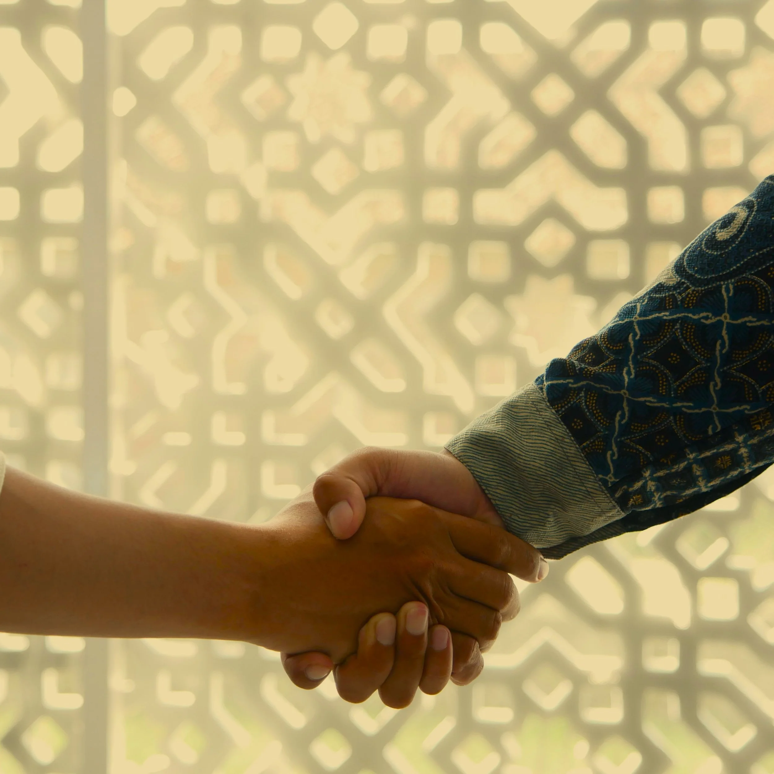 Two people shaking hands in front of a decorative window or screen with an intricate geometric pattern.