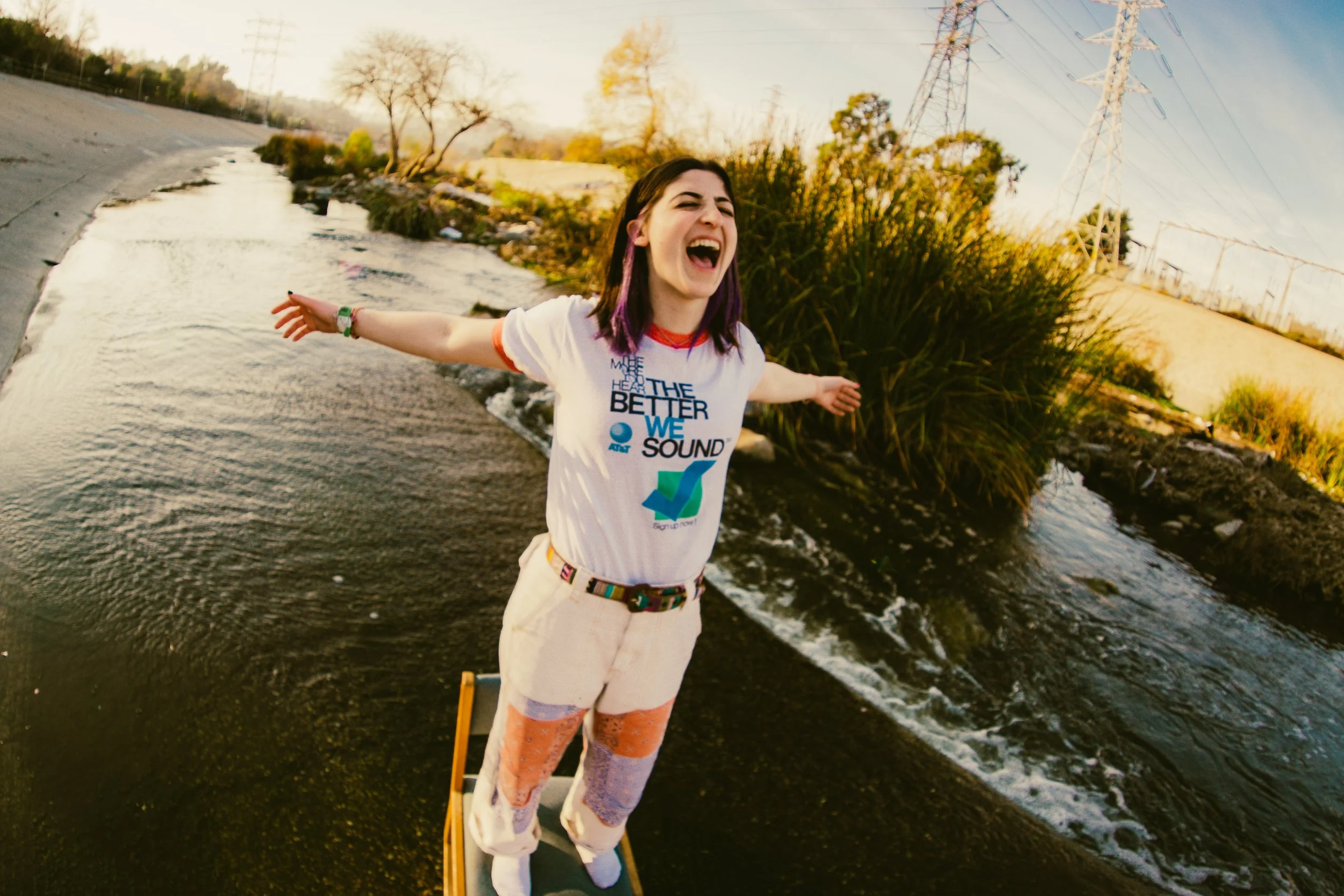 A woman standing on a longboard with arms outstretched near a creek, smiling with her mouth open, during late afternoon or early evening, with trees and power lines in the background.