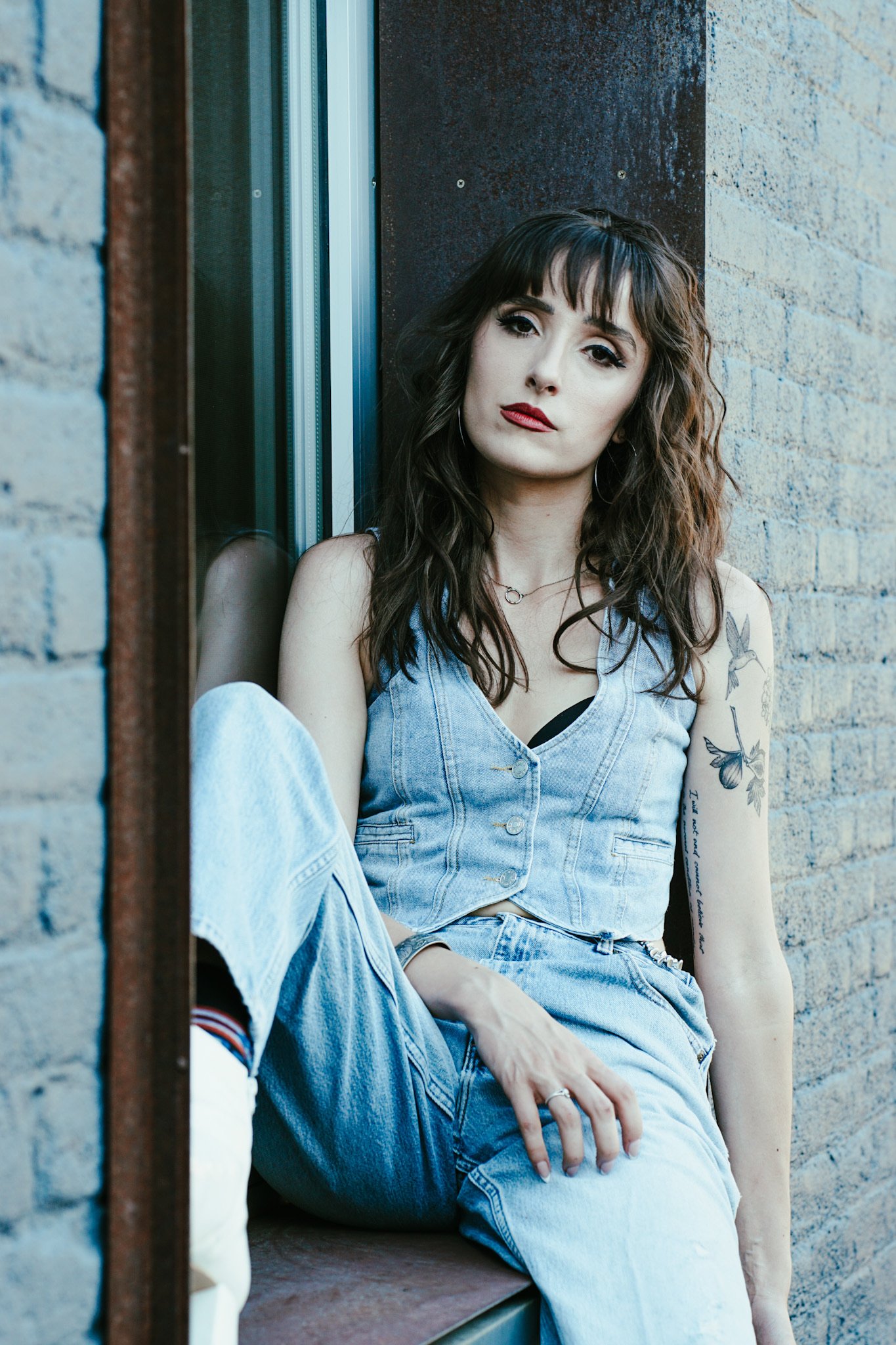 A woman with wavy brown hair and tattoos on her arm sitting on a ledge against a brick wall, looking at the camera.