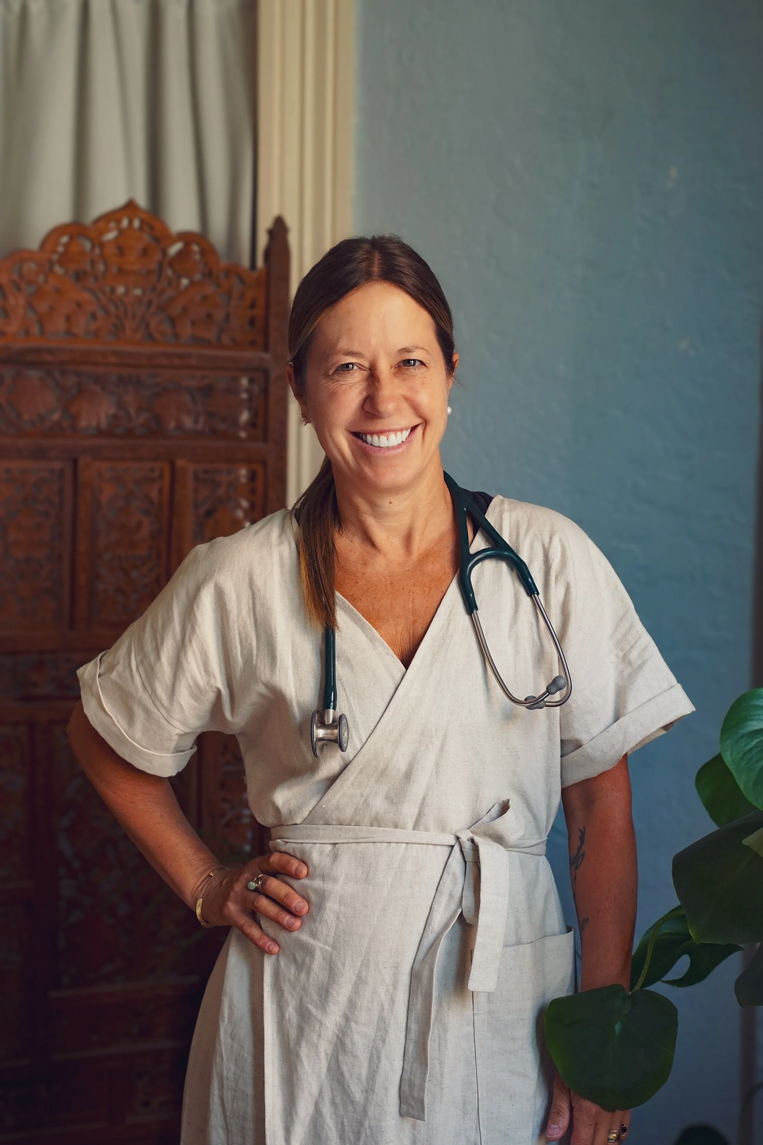 A smiling woman in medical scrubs with a stethoscope around her neck standing indoors, with a decorative wooden screen and a blue wall in the background, and a plant partially visible in the foreground.