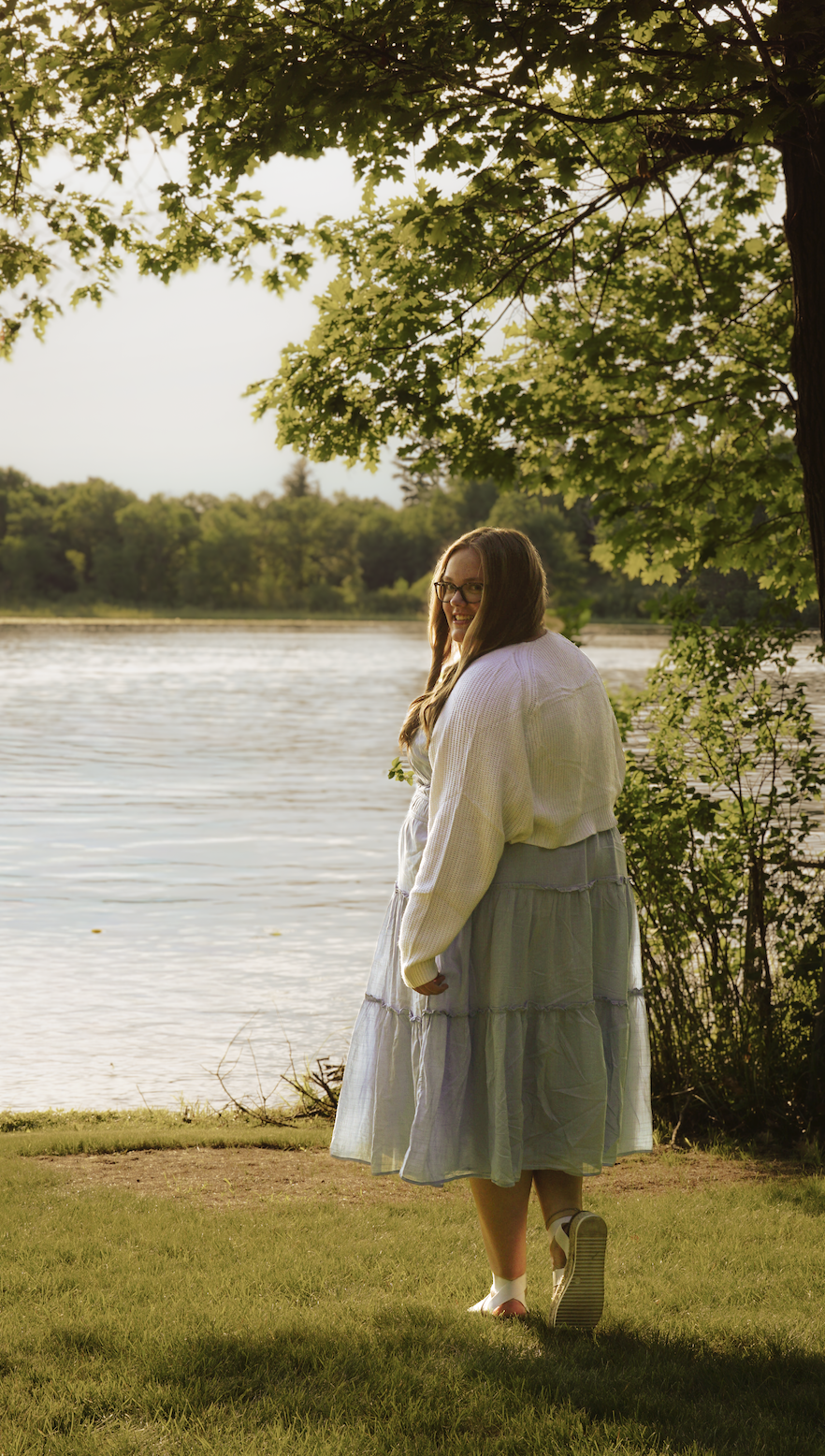 A young woman with glasses standing near a lake, smiling, under a tree with green leaves, during late afternoon.