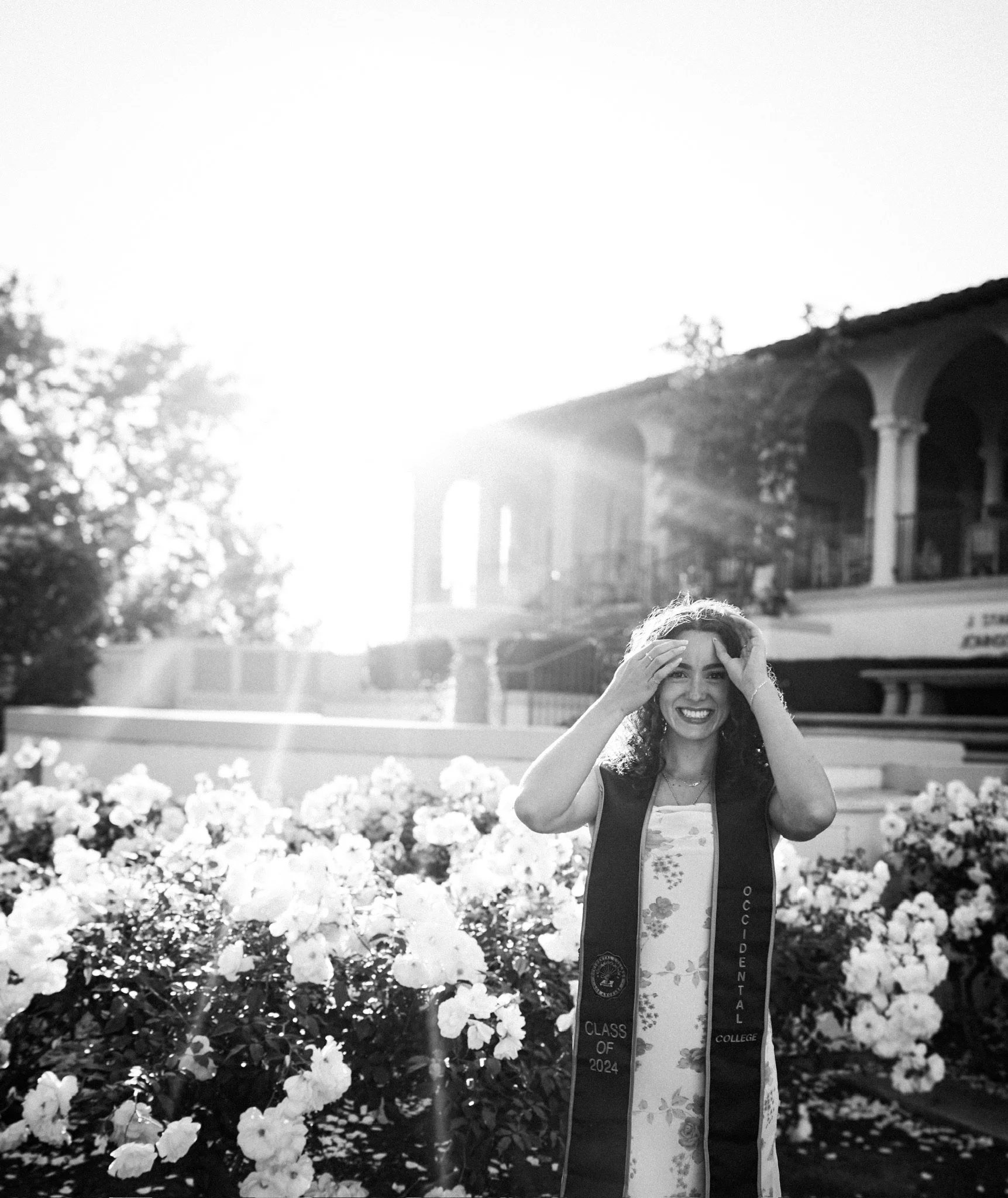 Smile of a woman standing outdoors with flowers and a bridge in the background, wearing a sash indicating her graduation class of 2024.