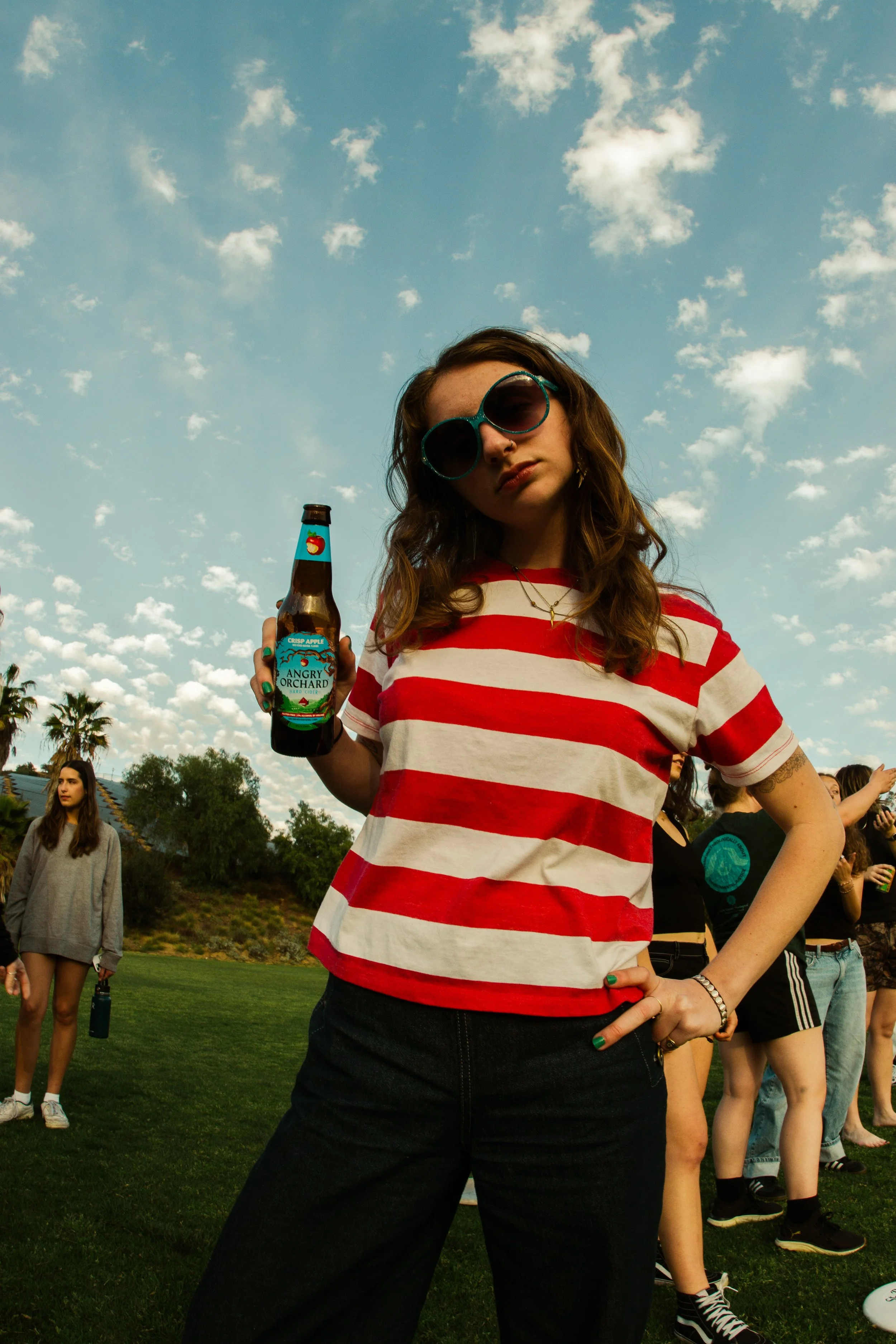 A young woman with wavy brown hair wearing large sunglasses, a red and white striped t-shirt, and dark jeans holding a brown glass bottle labeled 'Angry Orchard' with a blue and red design, outdoors with a group of people on a grassy field under a partly cloudy sky.