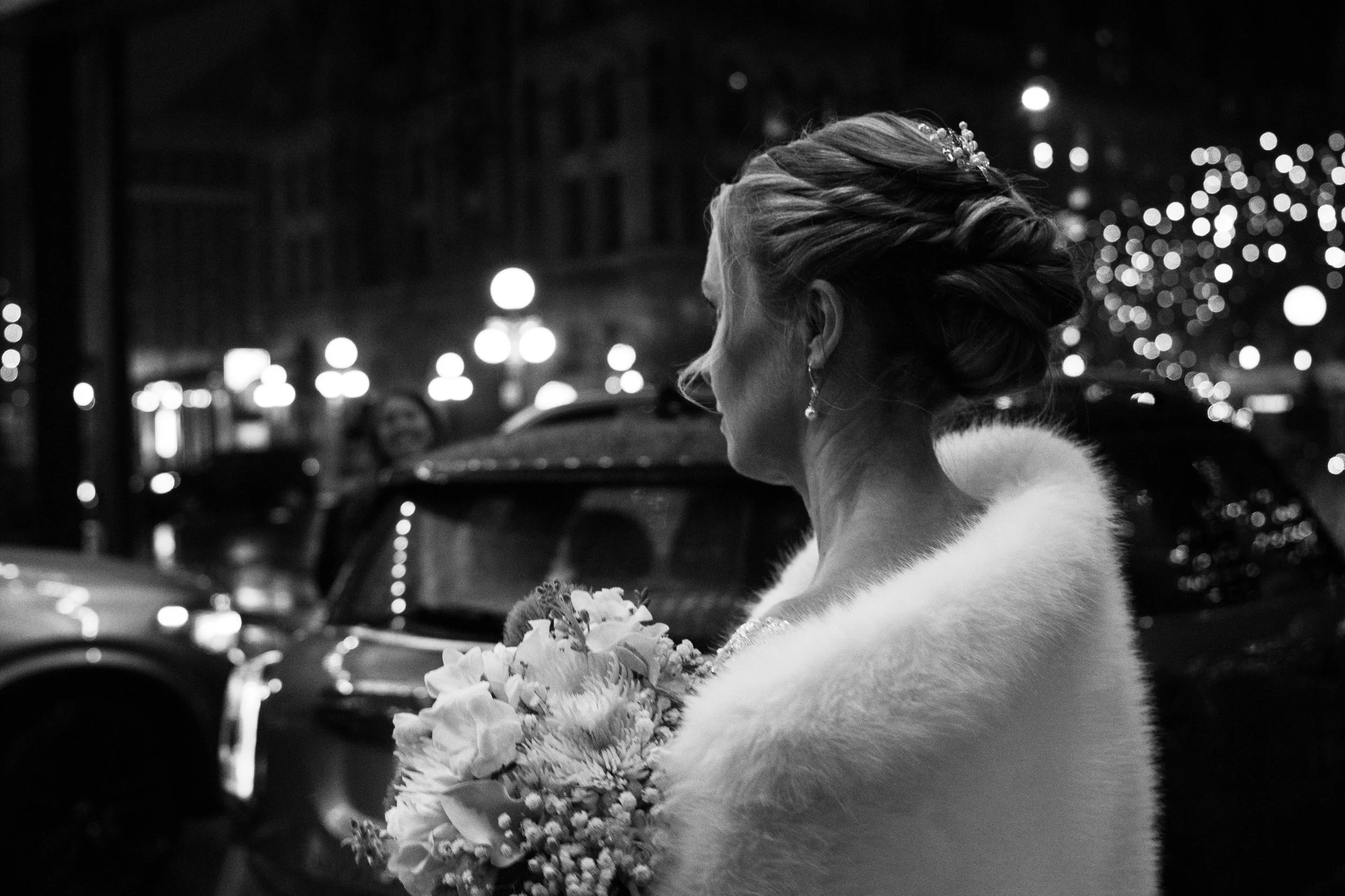 A woman with styled hair and earrings, holding a bouquet of flowers, standing outdoors at night with blurred city lights in the background.