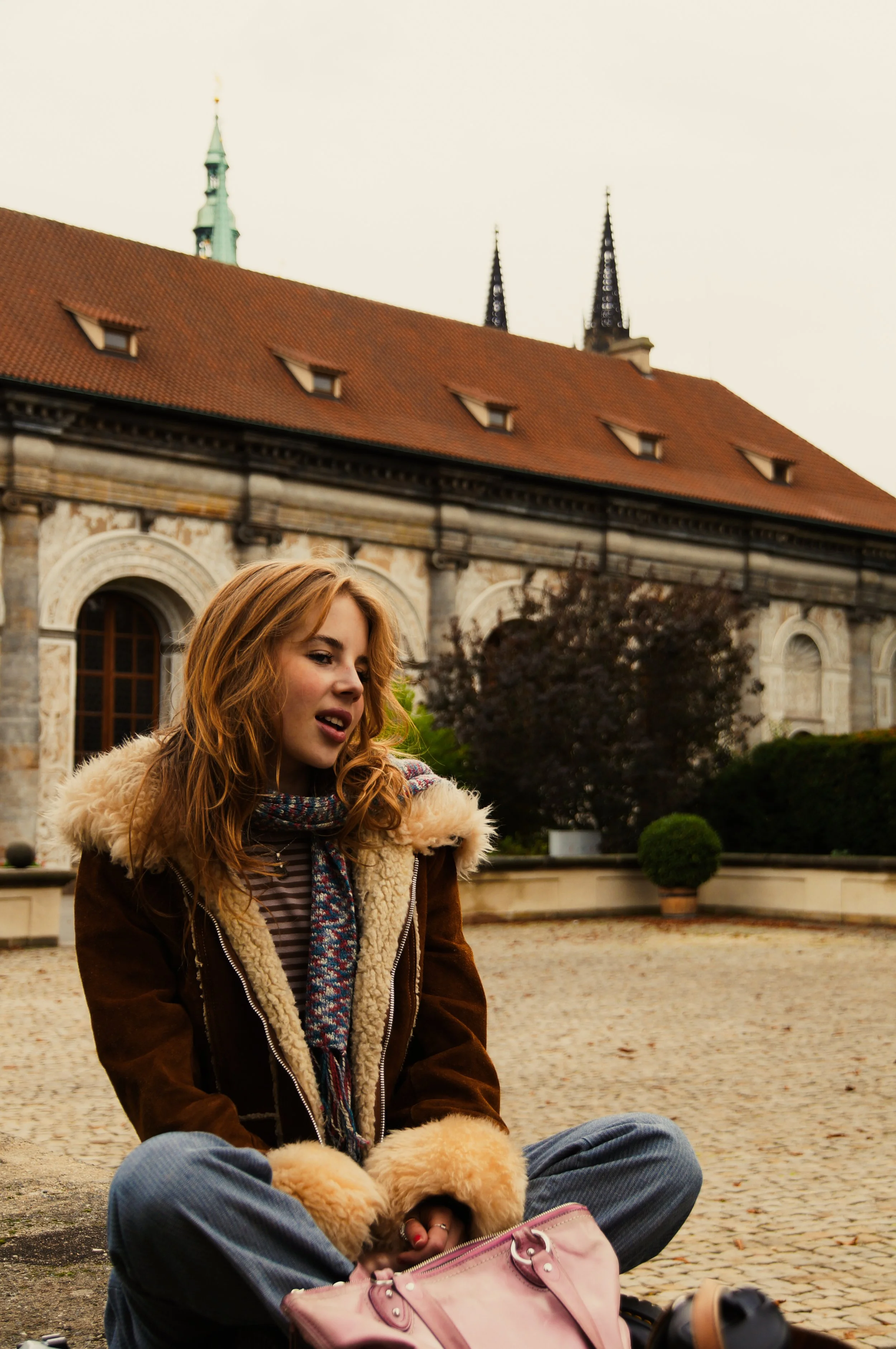 A young woman with red hair sitting on the ground, wearing a brown jacket with a fur collar, a striped shirt, and blue jeans. She is sitting in front of a historic stone building with a red-tiled roof and two towers with spires. She has a pink purse on her lap and is surrounded by fluffy puppies.