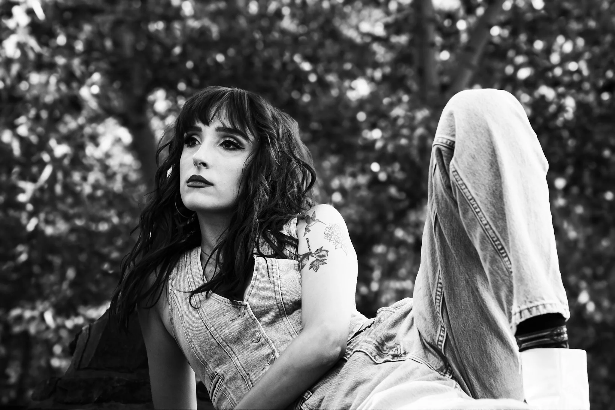 A black and white photo of a woman with shoulder-length wavy hair and tattoos on her left arm, sitting outdoors with trees in the background. She is wearing a sleeveless denim vest and looking to her left with a thoughtful expression.