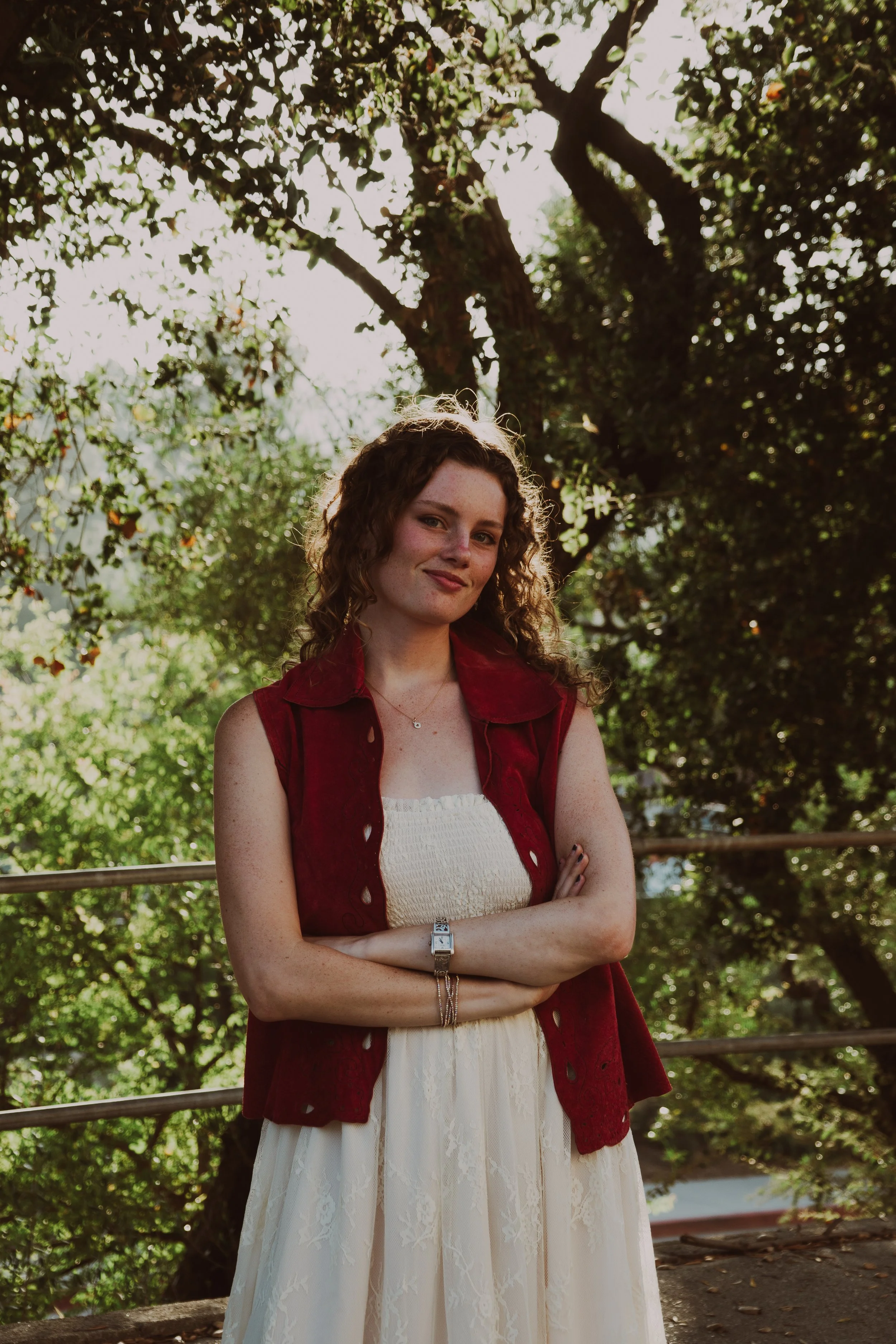 A woman with curly red hair standing outdoors in front of trees with green leaves. She is smiling, wearing a white dress and a red sleeveless vest, with arms crossed. The sunlight filters through the trees, creating a warm glow.