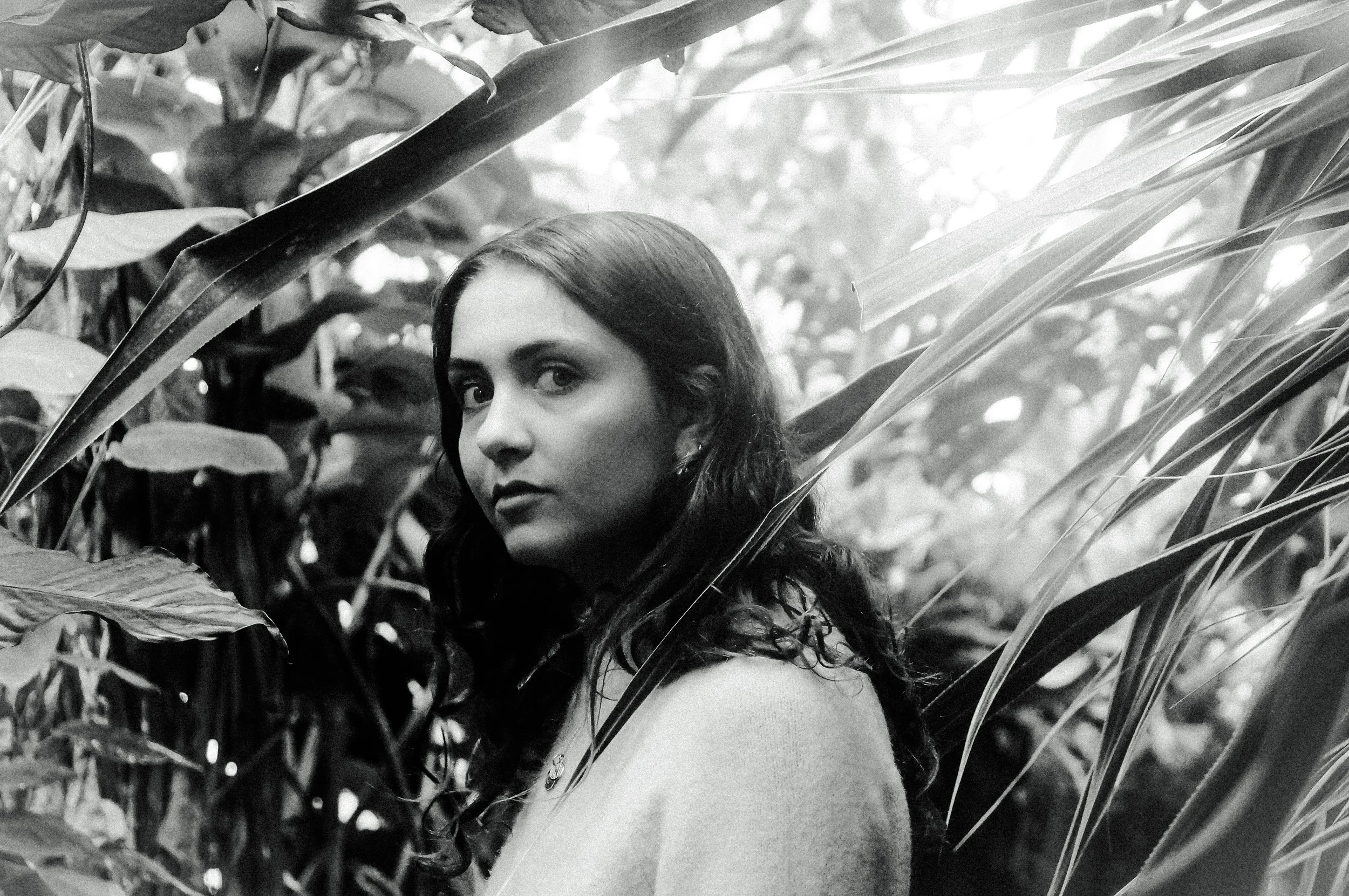 Black and white photo of a woman with long wavy hair among large tropical leaves, looking at the camera.