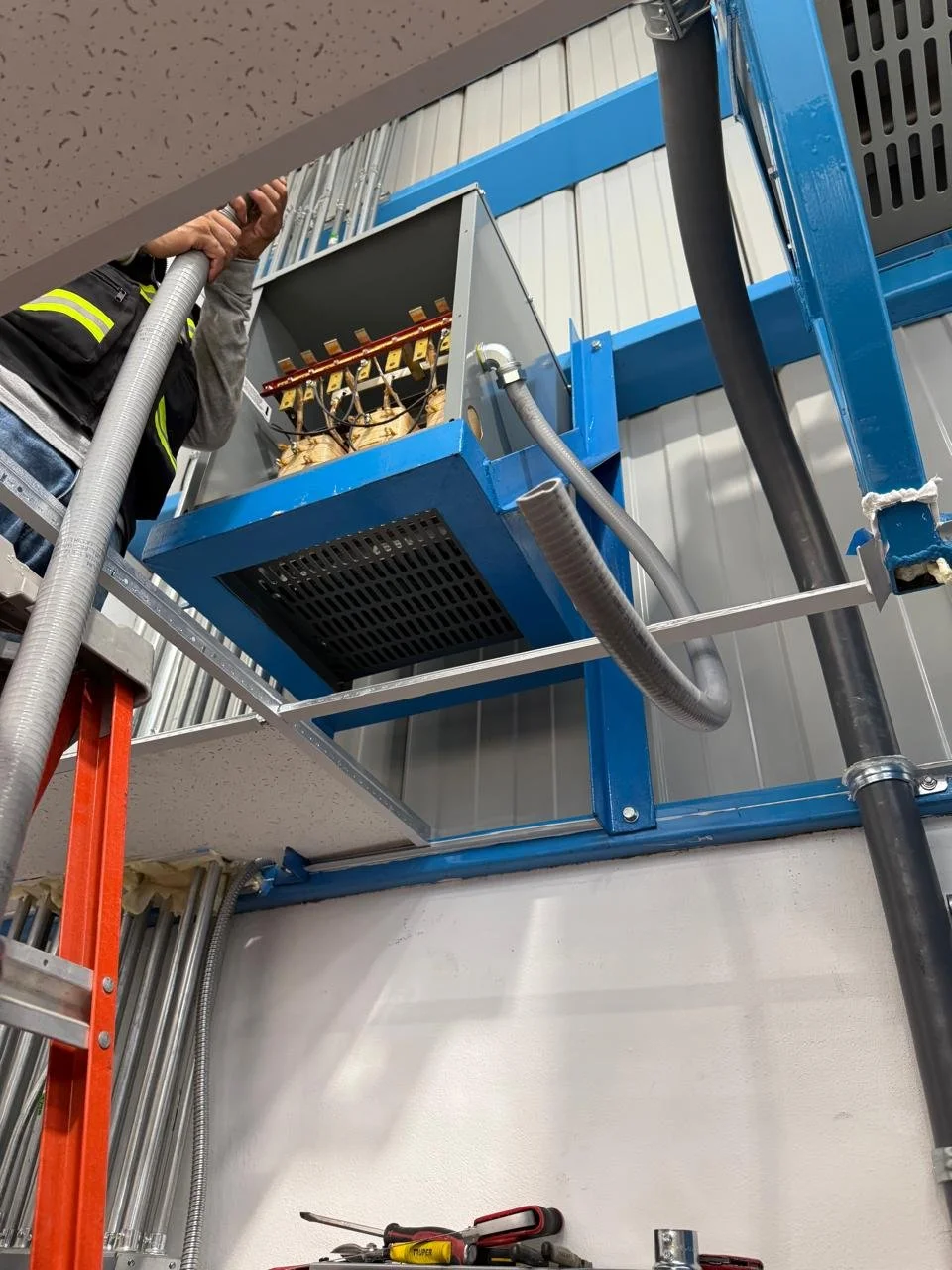 A worker on a ladder installing or maintaining electrical equipment in an industrial setting.
