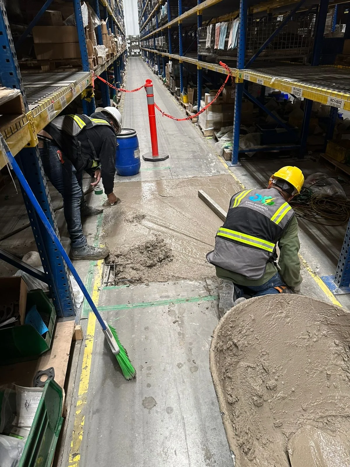 Construction workers repairing a damaged section of the warehouse floor, with one kneeling and one bending over, surrounded by construction tools, materials, and safety gear, with shelves stocked with boxes in the background.