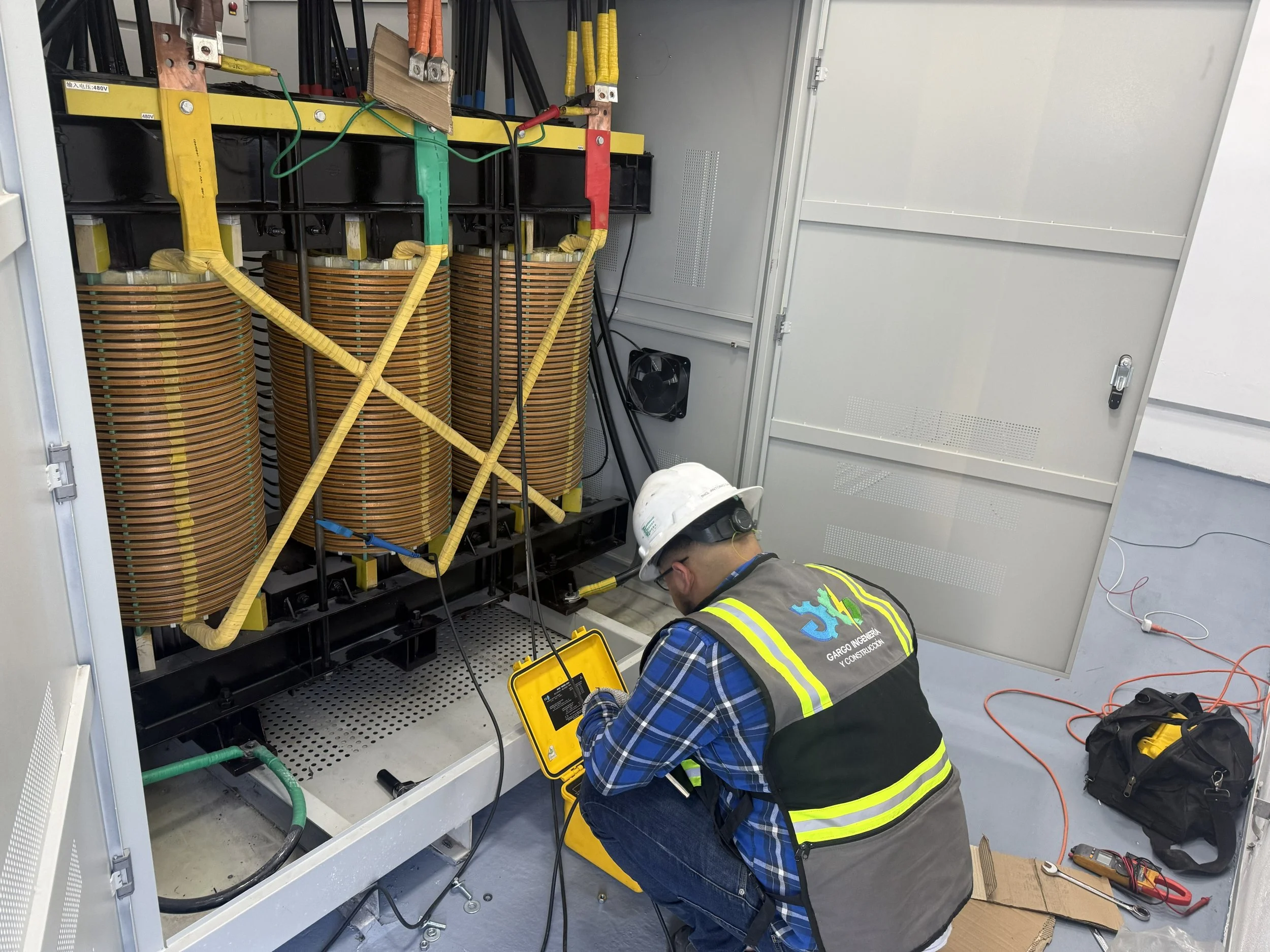 Worker in safety vest and helmet inspecting electrical components inside a large electrical cabinet with coils, wires, and testing equipment.