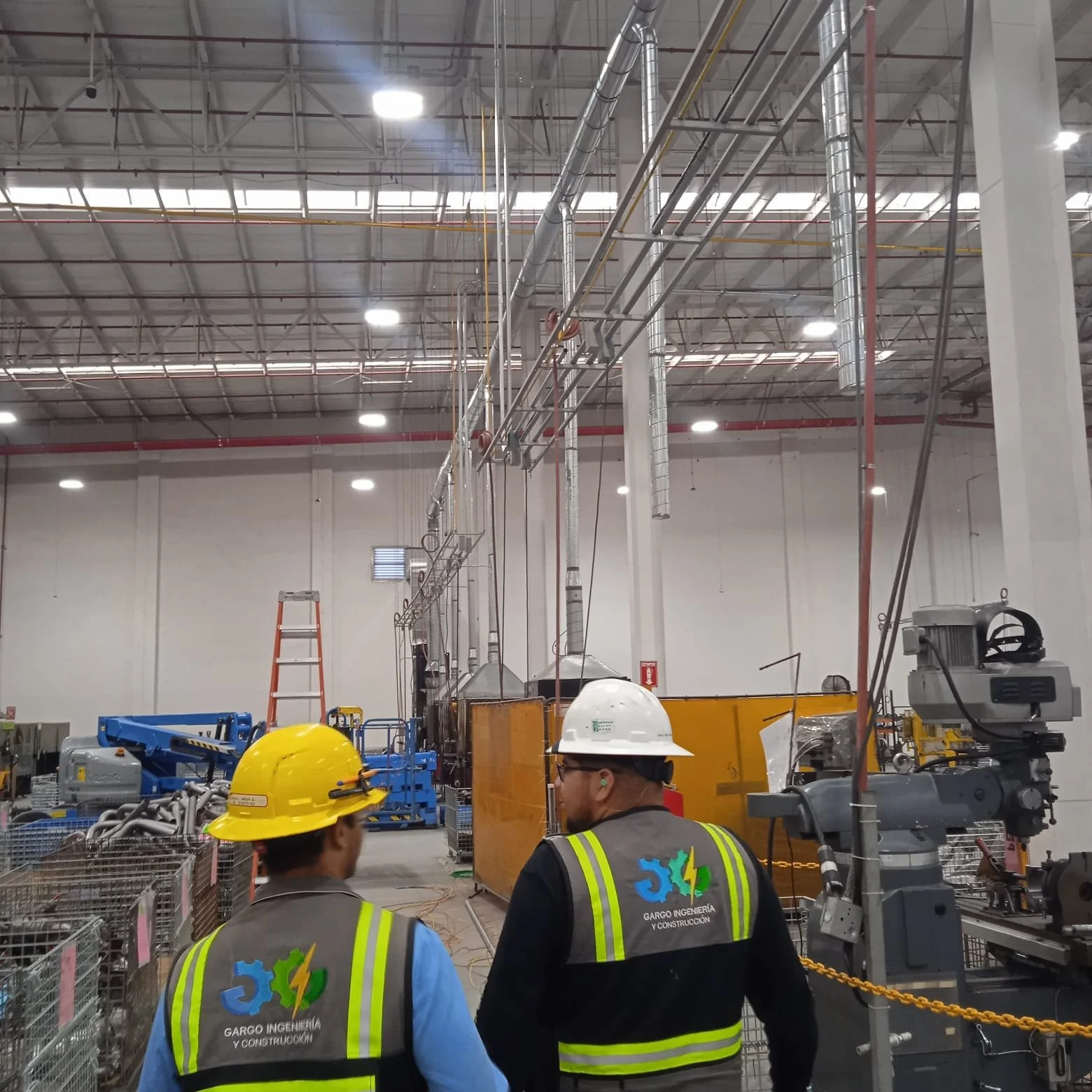 Construction workers in safety vests and helmets in an industrial facility with machinery, pipes, and scaffolding.