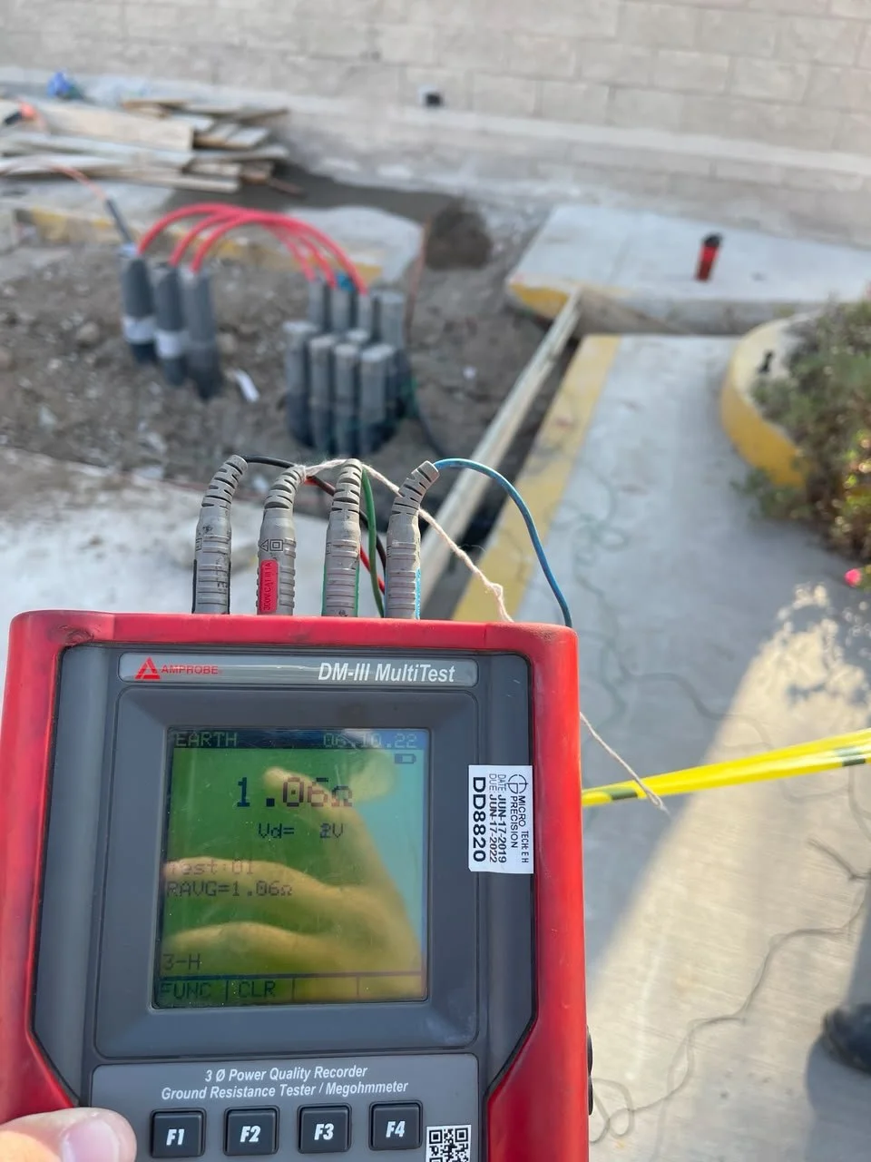 Electrician testing grounding resistance on construction site with gloves on, digital earth tester showing 1.06 ohms.