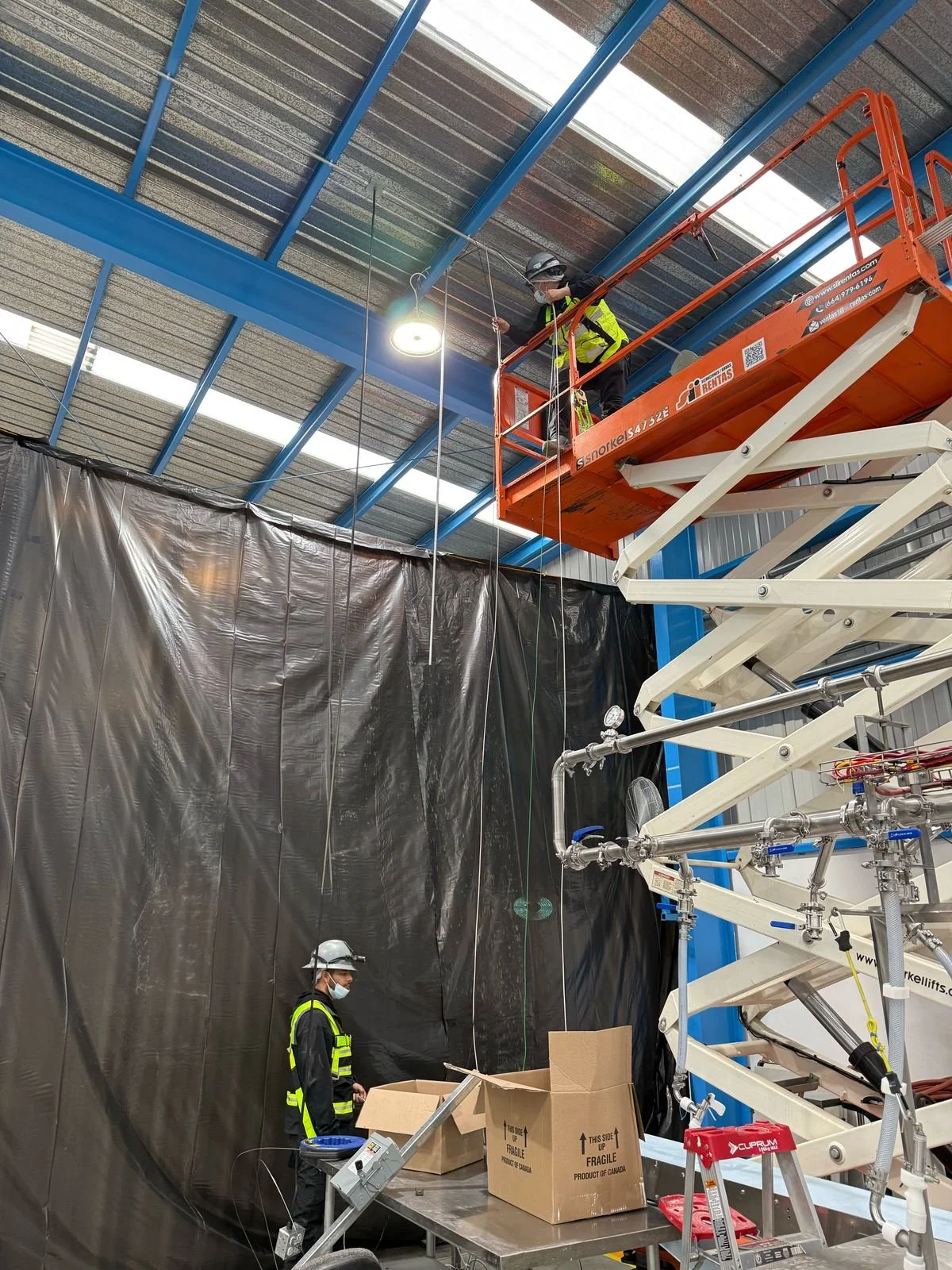Two workers in safety gear, including helmets and vests, operating in an industrial warehouse. They are working with a scissor lift and various equipment next to a large black curtain or tarp, with one worker on the lift and another on the ground nea