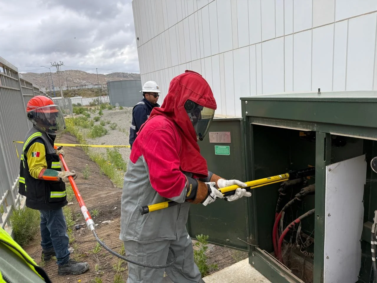 Workers in protective gear inspecting an electrical transformer outside a fenced industrial area, with cloudy sky and hills in the background.