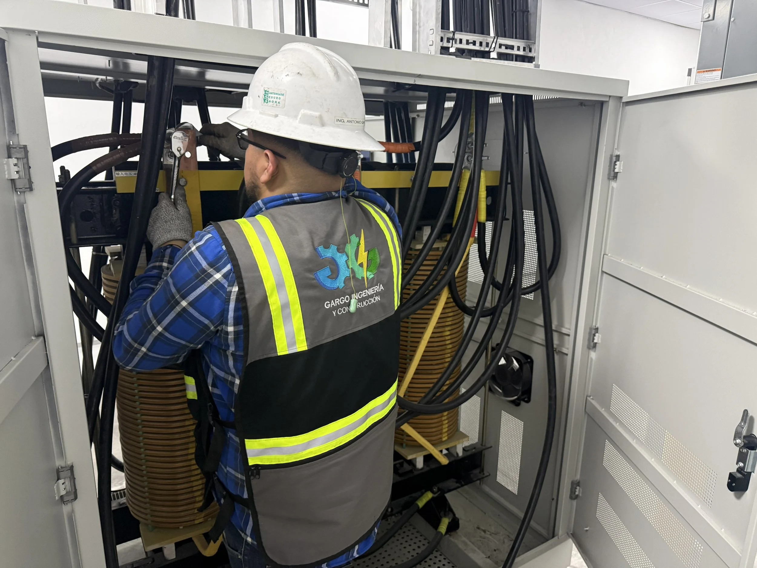 A technician wearing a white hard hat, safety glasses, and a high-visibility vest working inside an electrical control panel with many black cables and industrial equipment.