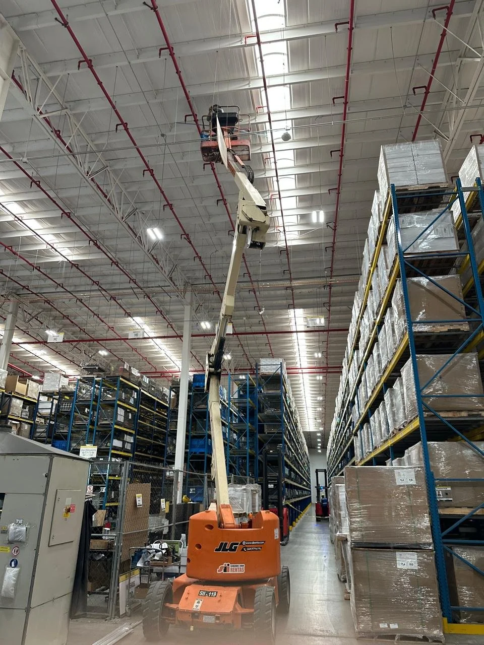 An industrial warehouse with high shelves filled with boxes and pallets. A boom lift is extended upward, reaching the ceiling to perform maintenance or installation work.