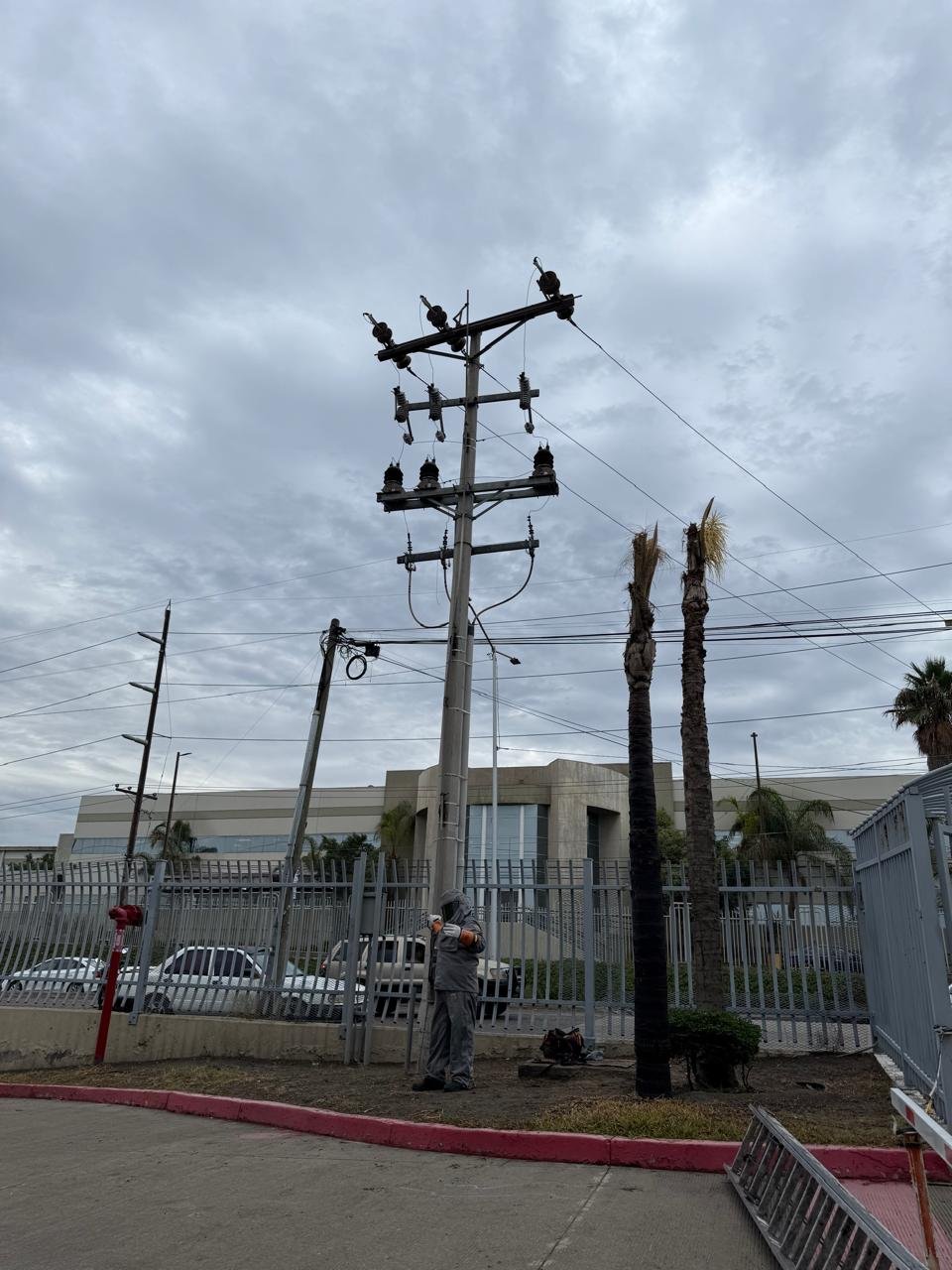 A utility worker in gray uniform standing next to a wooden utility pole with power lines and insulators, with a two-story building, a fence, and palm trees in the background under cloudy sky.