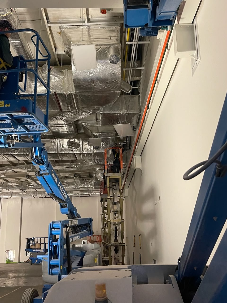 Construction workers on aerial lifts installing ductwork in an industrial space.