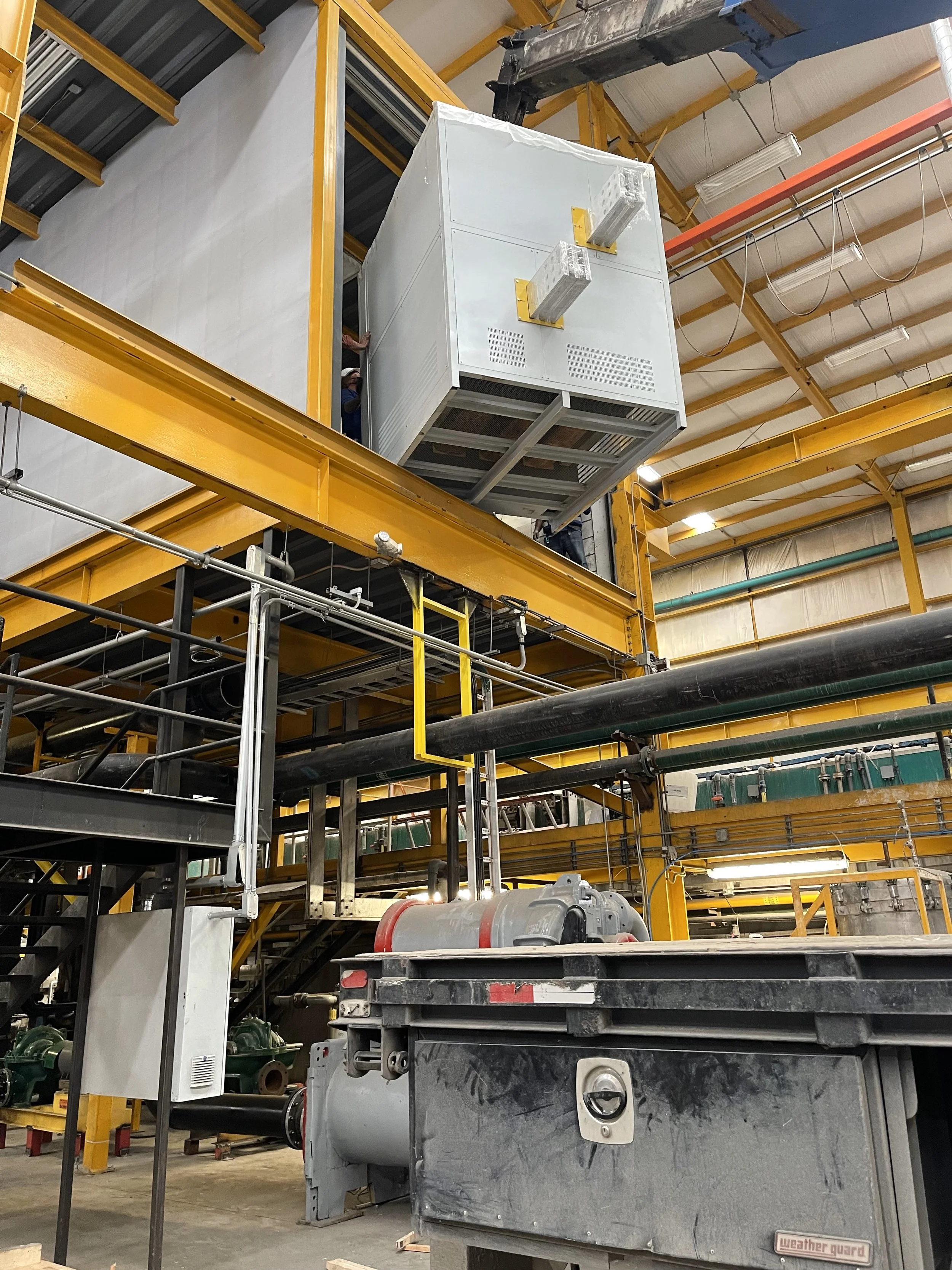 Industrial facility with yellow support beams and large gray machine suspended above a platform.
