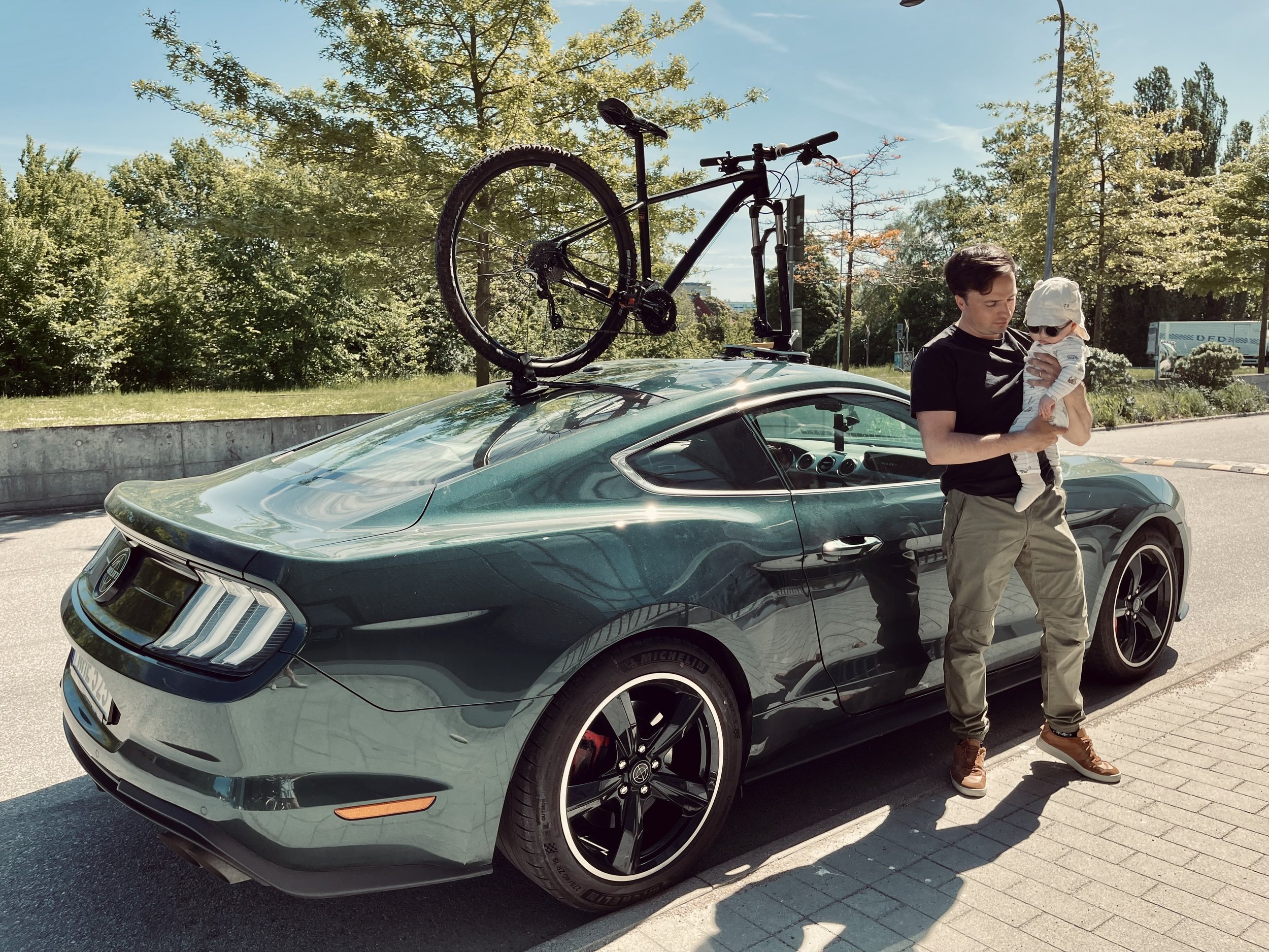 Timothy holding his son next to his Mustang Bullitt with a mountain bike mounted on the roof.