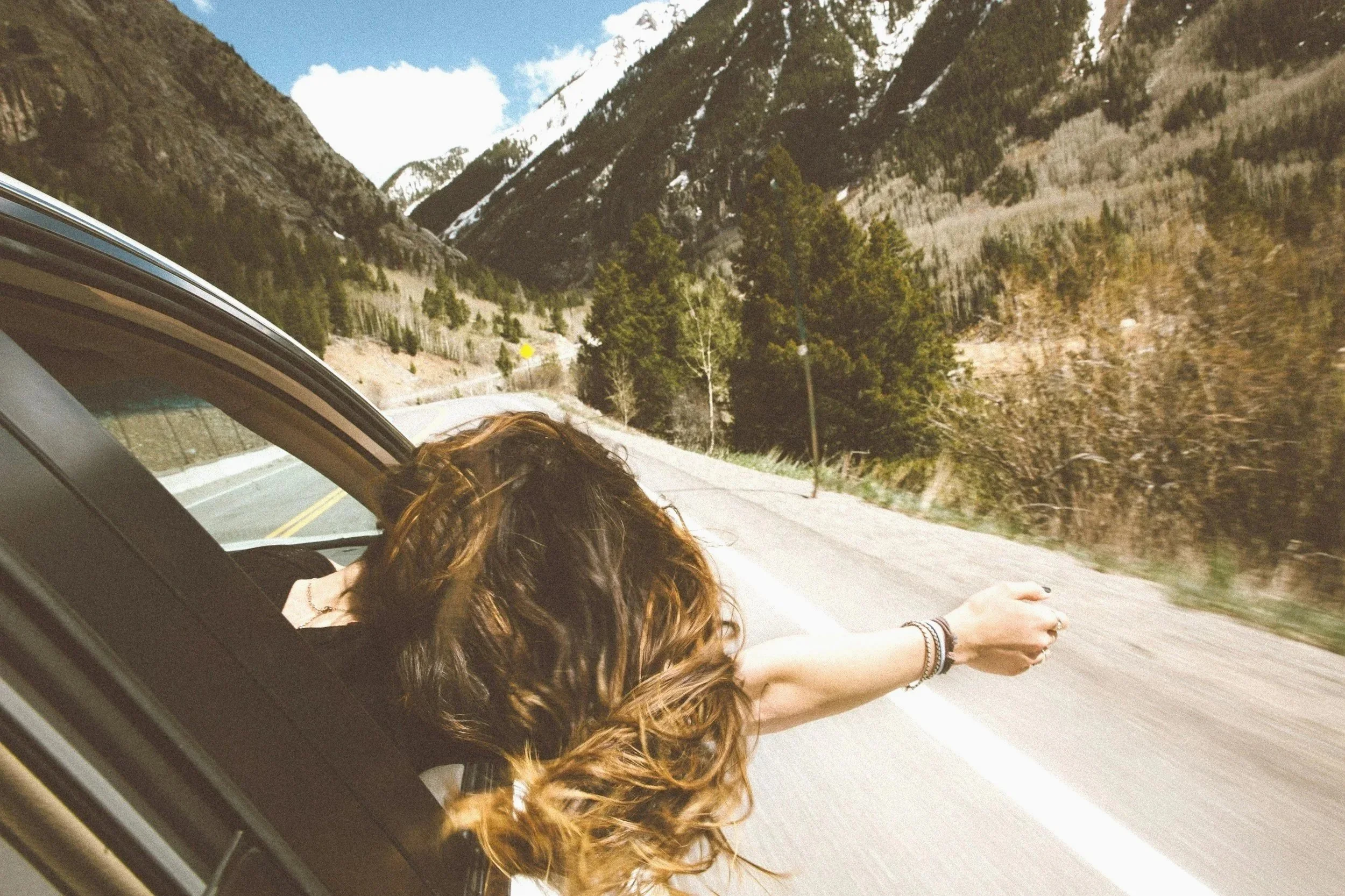 A person leaning out of a car window with arm extended outside, driving through a mountainous area with trees and snow-capped peaks in the background.