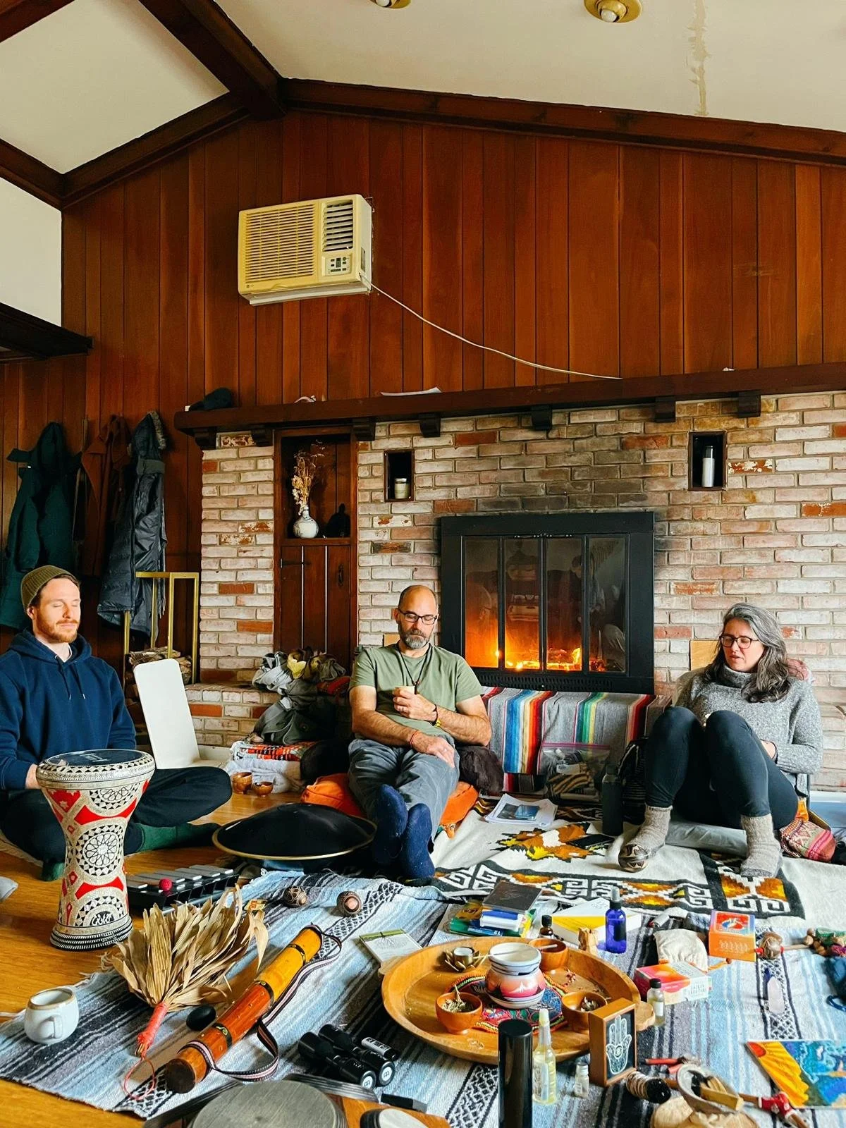 Three people sitting in front of a fireplace in a cozy living room, surrounded by various objects and decorations, with coats hanging on the wall.
