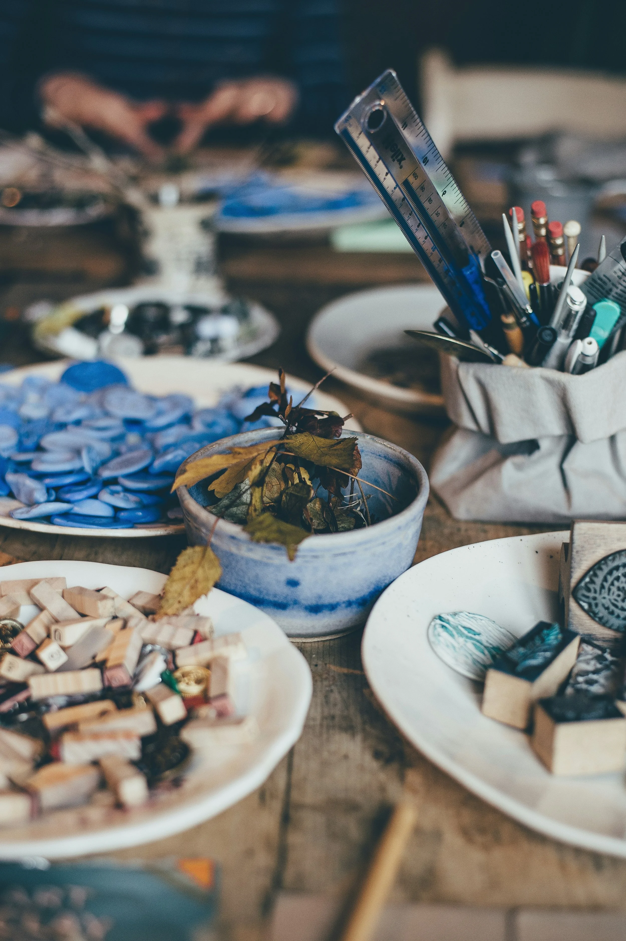 A table with various crafting supplies, including bowls of small tiles, pieces of broken ceramics, and tools like a ruler and pens, with a blurred background of artistic workspace.