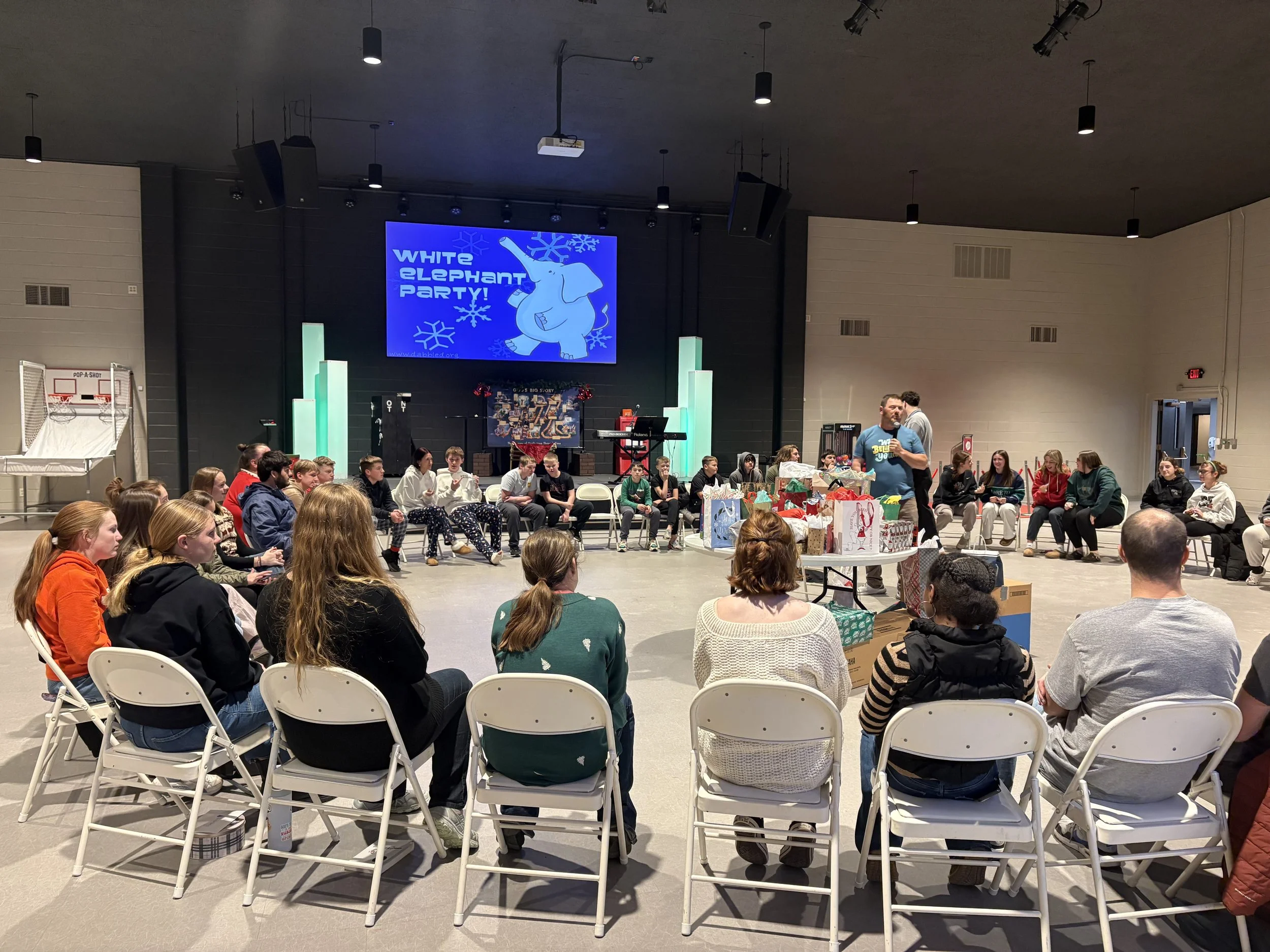 A group of middle school and high school students, facing a stage with a large blue screen that says 'White Elephant Party!' with a cartoon elephant and snowflakes. We have many great activities for students, teenagers, and kids at church.