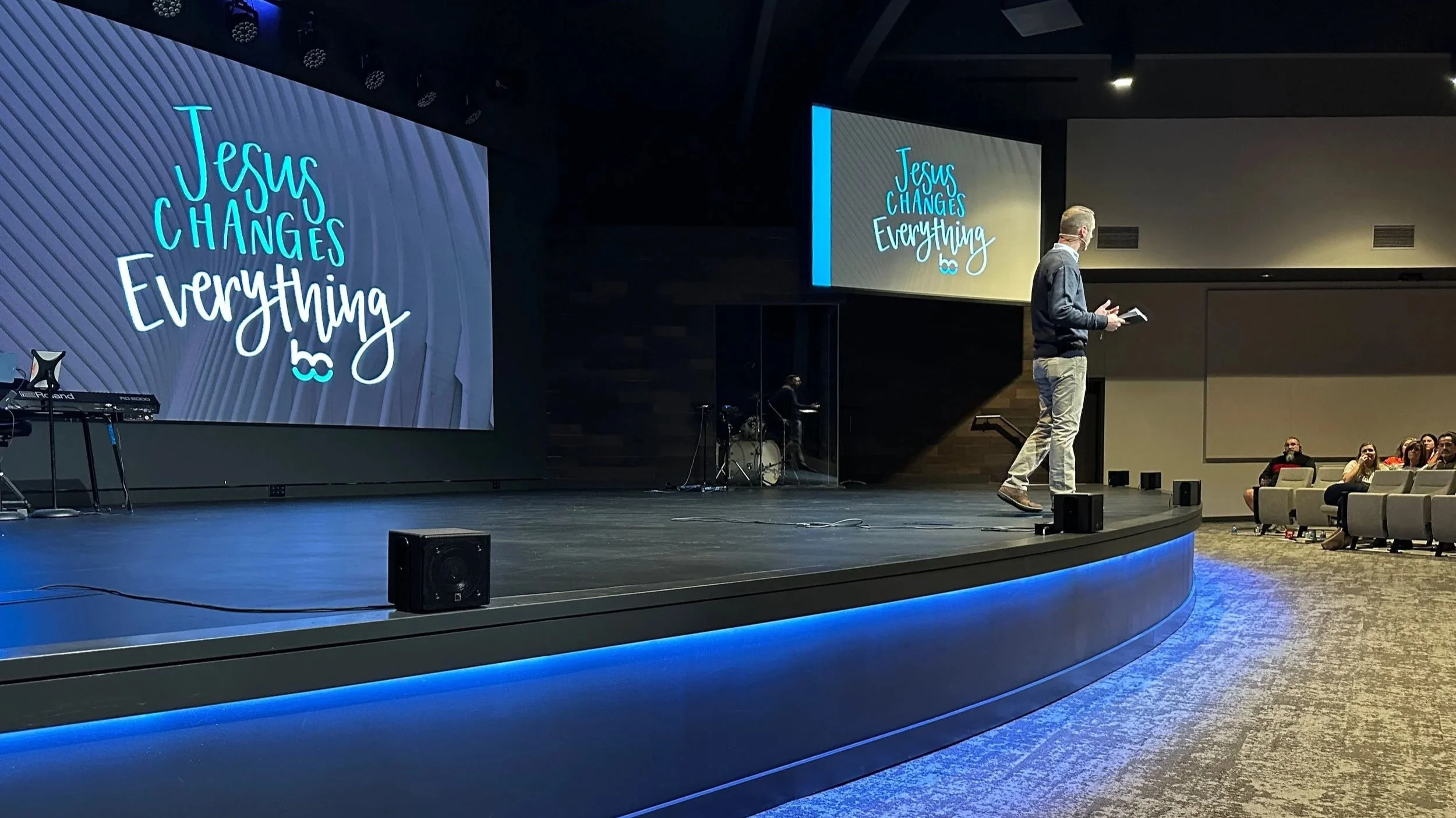 A man standing on a stage presenting a Bible based Sermon from the words of Jesus. Two large screens display the message 'Jesus Changes Everything' in colorful text. There are a many audience members seated and watching.