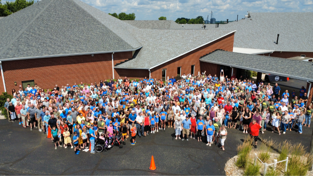 A large group photo after church as they are gathered outside a brick building with a gray roof, on a sunny day.