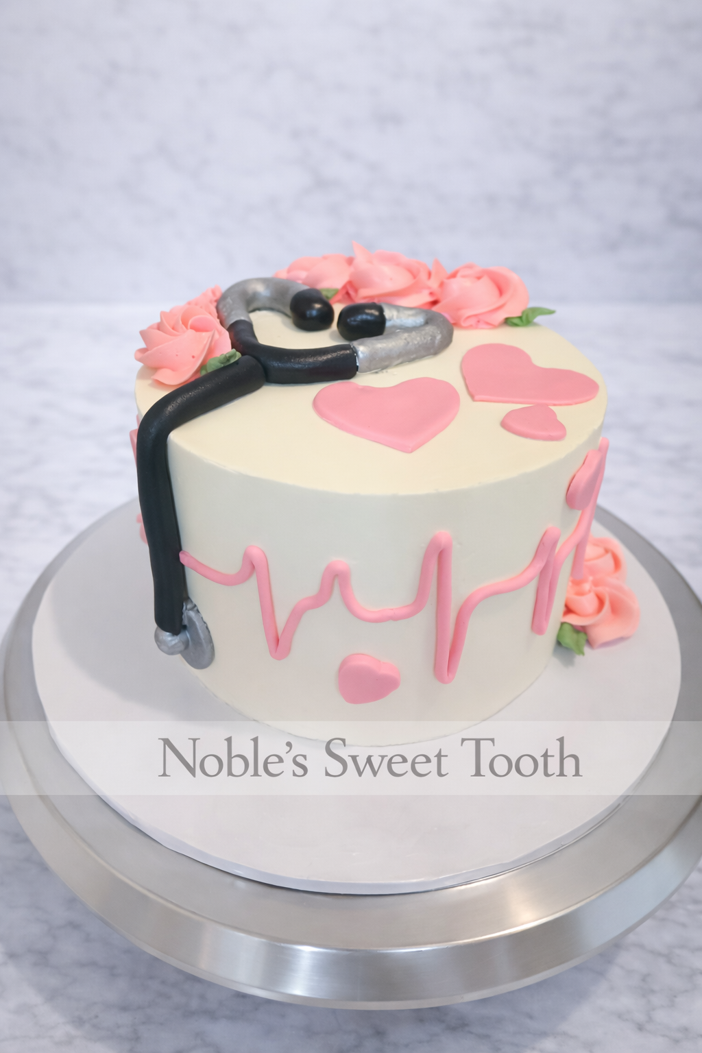 A round cake decorated with a stethoscope, pink roses, and pink heart shapes, with a cardiology-themed design and the words 'Noble's Sweet Tooth' on the front.