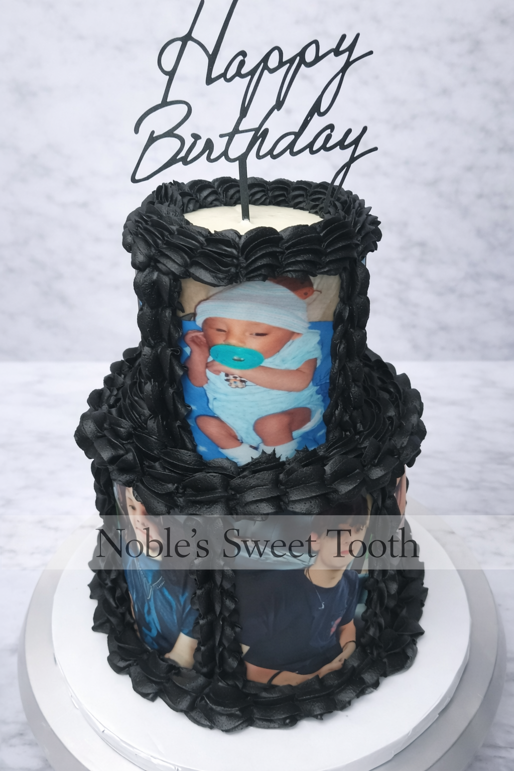 A birthday cake decorated with black frosting and photos of a young boy, topped with a 'Happy Birthday' cake topper. The cake has a label 'Noble's Sweet Tooth'.