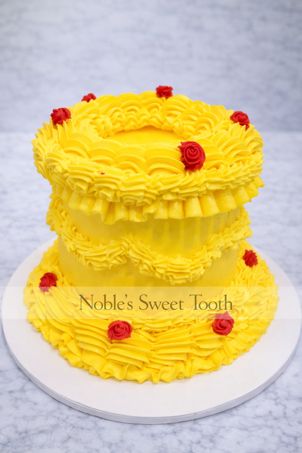 A yellow, two-tiered cake decorated with red roses and piped frosting, with a label that reads "Noble's Sweet Tooth."