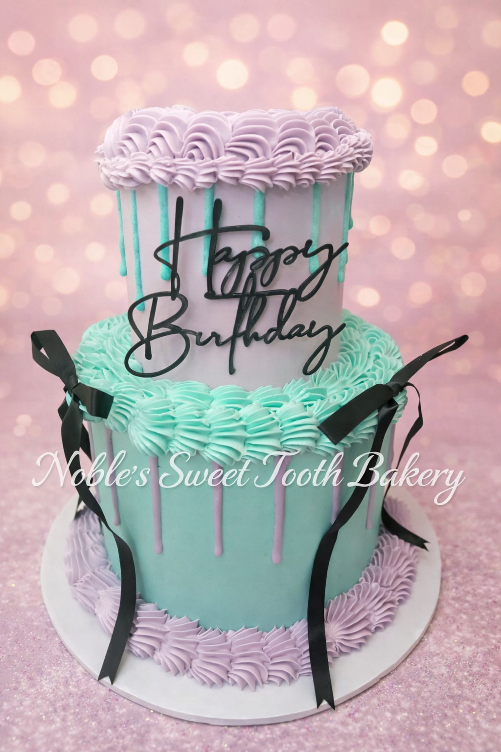 A two-tier birthday cake with pastel pink and mint green frosting, decorated with black ribbons, pink and purple piped frosting, and a "Happy Birthday" message written in black cursive. The cake sits on a white base with a pink, glittery background.